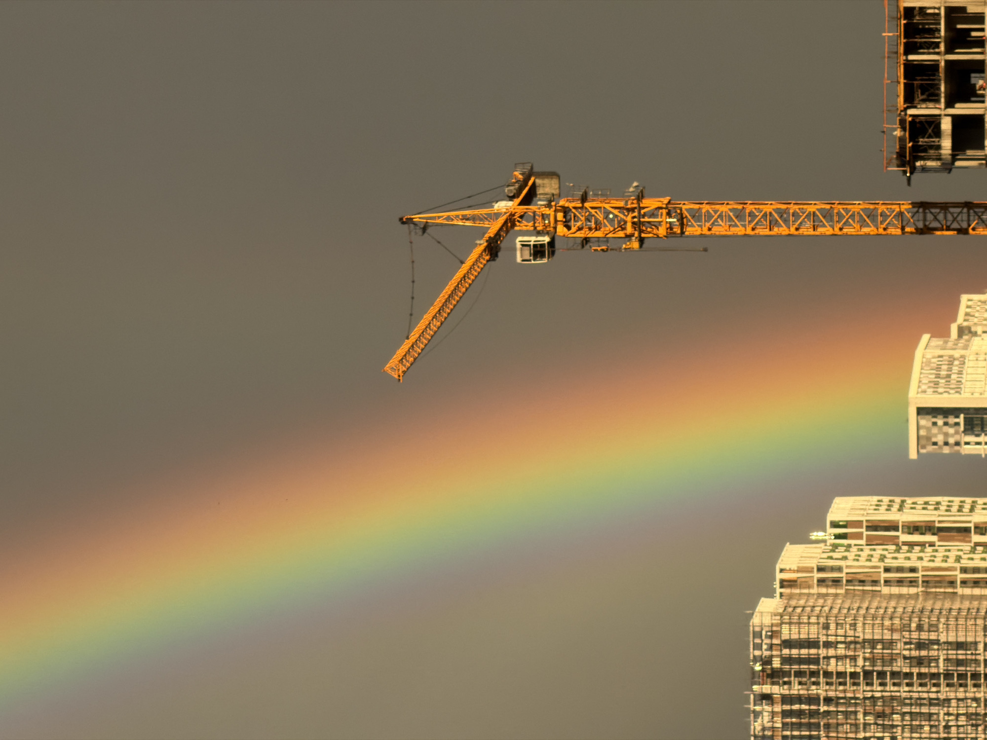 The image captures a construction site in Izhevsk, Russia, under a gray, overcast sky, with a vibrant rainbow arching across the left side of the frame. The rainbow displays the full spectrum of colors, from red on top to violet at the bottom, creating a striking contrast with the muted background. Prominently featured is a tall, yellow construction crane positioned in the mid-ground to the right. Its articulated arm extends upwards and to the left, appearing to hover over the scene. In the left mid-ground, several unfinished high-rise buildings are visible, their skeletal structures indicating ongoing construction. One building is particularly detailed, with a facade of numerous windows and balconies. To the far right, another construction project is partially visible, showing concrete pillars and a basic framework. The overall scene depicts urban development against a backdrop of a natural phenomenon.