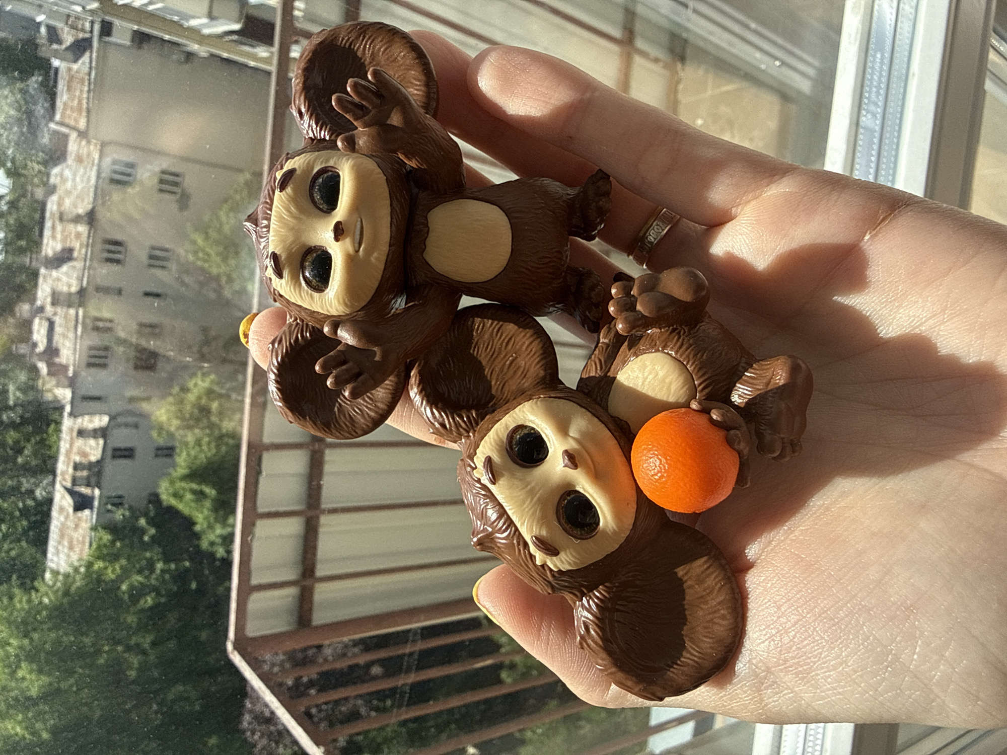 A pale hand holds two Cheburashka figurines. The top figurine is positioned upside down. The bottom Cheburashka holds a small orange ball. A ring is visible on one of the fingers. The background shows a view through a window: buildings and trees are visible. The location is Moscow, Russia.