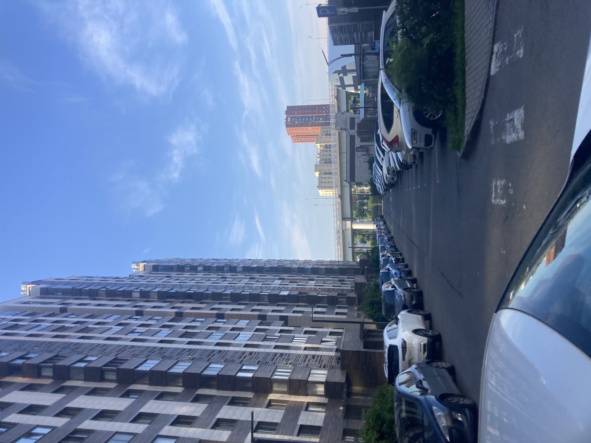The image captures a street view in Moscow, Russia, likely during daytime, under a clear blue sky with scattered wispy clouds. The scene is dominated by a modern, tall residential building with a facade of alternating dark brown and light-colored panels, featuring numerous windows. To the right, a row of parked cars lines the asphalt road. In the background, an elevated highway or bridge structure crosses the scene, with other buildings visible beyond it, including a prominent reddish-orange multi-story building and a lighter-colored, lower-rise building. The environment appears to be an urban residential area. No people are visible in the image, and no actions or interactions are depicted.