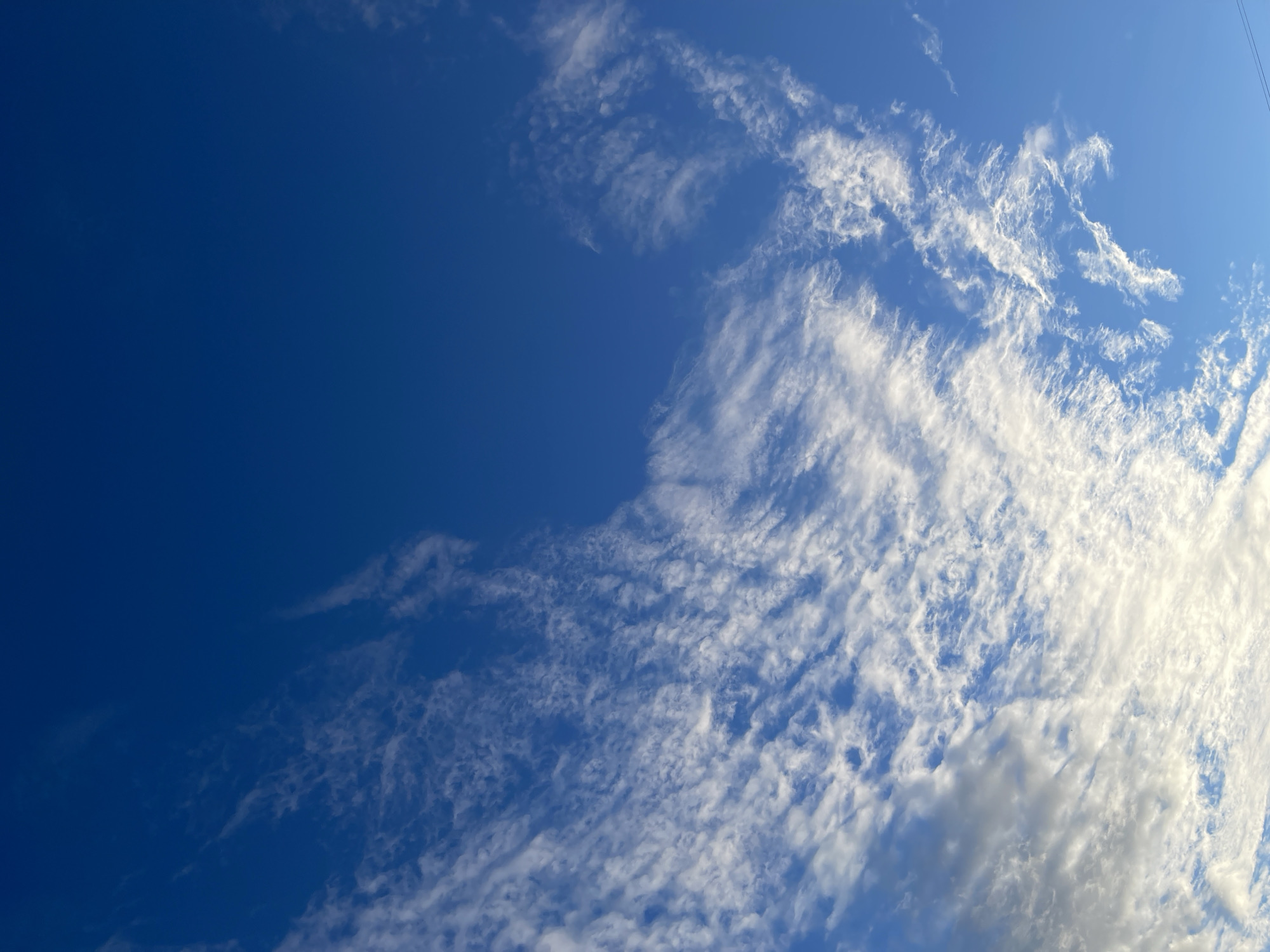 The image depicts a vast expanse of a deep blue sky, sparsely populated with wispy white clouds. The clouds are scattered and appear to be moving across the sky, with some areas showing denser formations and others almost completely clear blue. The lighting suggests it is daytime, with sunlight illuminating the clouds from above and to the right, creating bright highlights and softer shadows. There are no discernible objects, people, or text visible in the frame. The scene evokes a sense of openness and natural beauty, typical of a clear day with some atmospheric activity. The location context provided is Moscow, Russia, which implies a temperate climate where such sky conditions are common.
