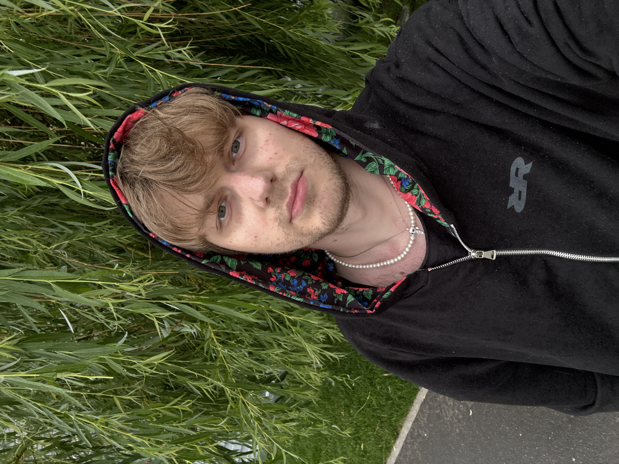 A young man with blonde hair and light-colored eyes is taking a selfie outdoors. He is wearing a black zip-up hoodie with a floral pattern lining the hood. He has a pearl necklace on. The man is positioned in the lower right of the frame, with his head tilted slightly to his left. Behind him and to his left is a dense curtain of lush green foliage, likely willow branches. The ground in the foreground is a mix of grass and asphalt. The lighting suggests it is daytime, with no strong shadows indicating a time of day. The overall mood is relaxed and casual. The hoodie has a subtle gray logo on the right chest area, which appears to be stylized letters.