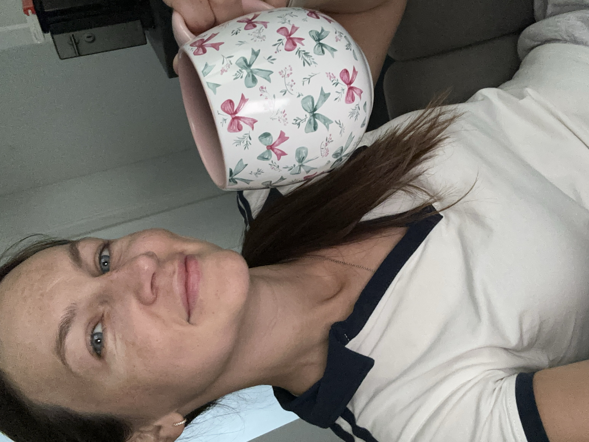 A woman with brown hair and blue eyes holds a mug with a bow pattern. She is wearing a white polo shirt with dark blue trim. The background is blurry and appears to be indoors, possibly a living room. The image was taken in Kutenholz, Germany.