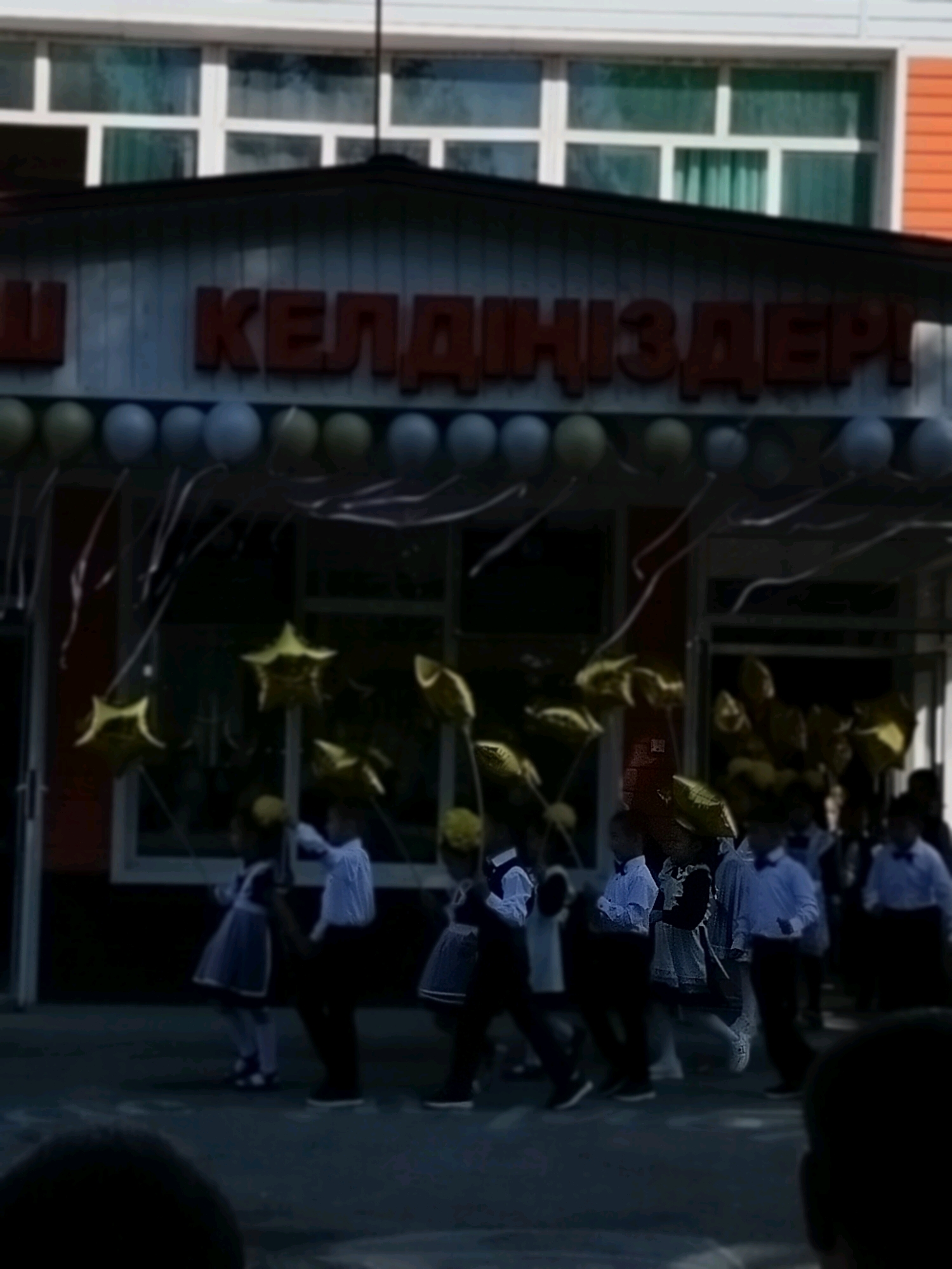 The image depicts an outdoor scene likely in the city of Kölkent, Kazakhstan, characterized by a festive atmosphere. In the background, a building with large windows and orange siding is visible, with a dark awning above the entrance. The awning is decorated with balloons in varying shades of grey and green, and streamers hang from it. A large red sign above the awning reads "Ш КЕЛДІҢІЗДЕР!", which translates to "Welcome!" in Kazakh.
In the foreground, a group of children, dressed in what appears to be school uniforms, are the primary subjects. Some are wearing dark dresses with white collars and cuffs, while others are in light-colored shirts and dark pants or skirts. Many of the children are holding gold star-shaped balloons. They appear to be engaged in some form of activity, possibly a procession or a dance, as they are positioned in a line and some are looking towards the viewer. The ground appears to be asphalt or pavement. The overall lighting suggests it is daytime, though the image has a slightly dark and possibly overcast quality.