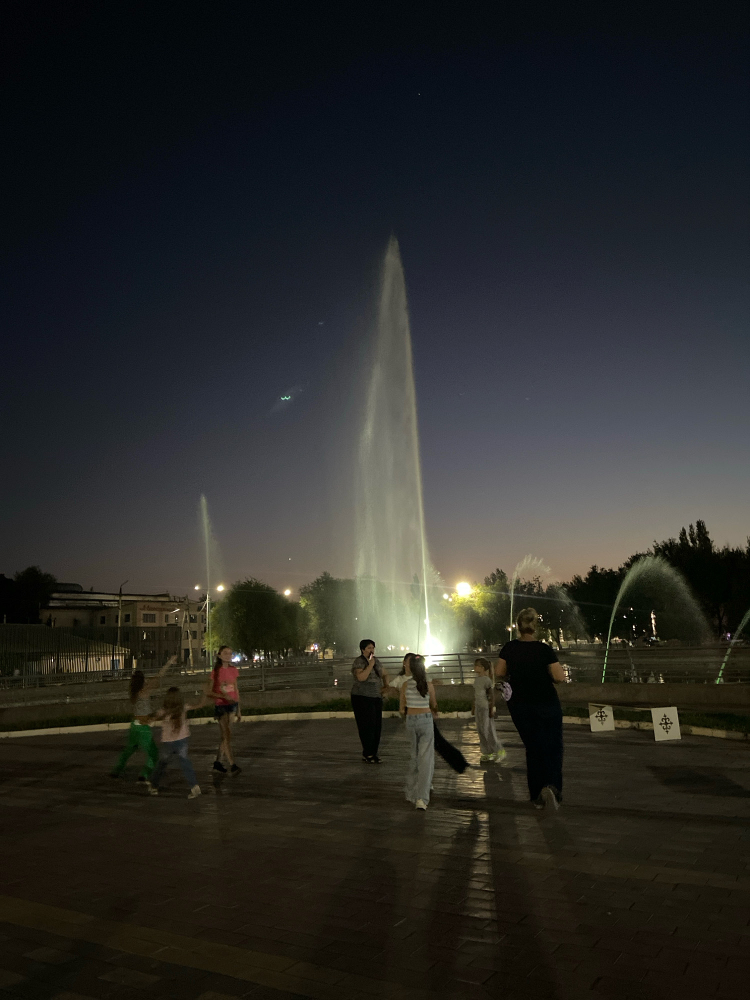 In Shymkent, Kazakhstan, at night, several people are gathered near a large illuminated fountain. The sky is a deep twilight blue, with faint streaks of light visible. The fountain's powerful spray creates a tall, luminous plume, reflecting the surrounding lights. In the foreground, a group of individuals, including children, are walking and interacting. To the left, a building with lights is visible, and trees line the background. The ground appears wet, possibly from the fountain's mist. The overall scene depicts a lively public space in the evening.