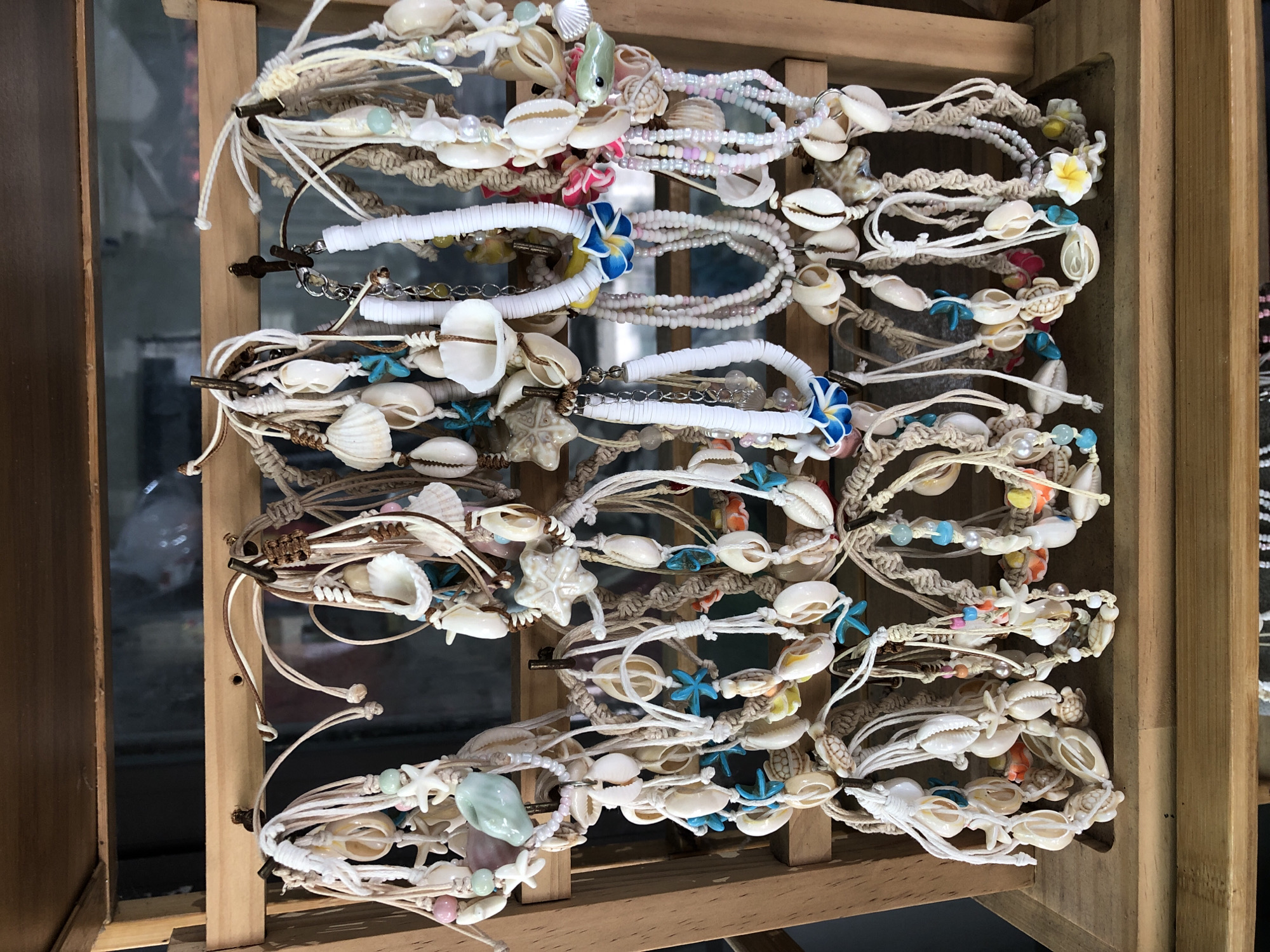 This media file shows a close-up, top-down view of an assortment of seashell and beaded bracelets displayed on a wooden rack. The bracelets are arranged in several rows, predominantly in shades of white, beige, and light blue, with accents of pink, green, and yellow. Many feature small shells, pearls, and colorful beads. Some bracelets are adorned with decorative elements like small starfish and flowers. The setting appears to be a market or retail stall, likely in a tropical or coastal location, given the theme of the jewelry. The lighting suggests it could be daytime, with natural light filtering in. The overall impression is of handcrafted, beach-themed accessories being offered for sale. There is no visible text in the image. The location context is Đà Nẵng, Vietnam, which aligns with the type of souvenirs often found in such areas.