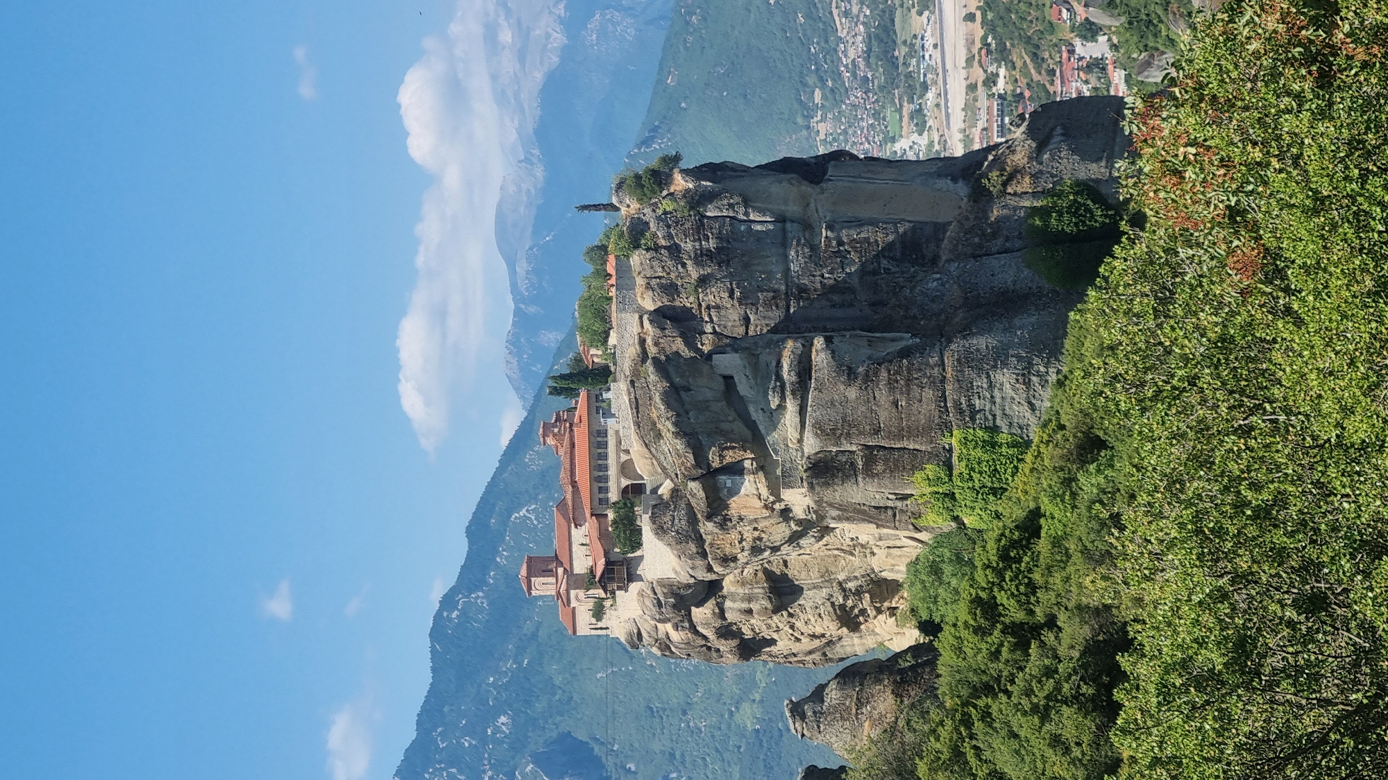 The image shows the Meteora monasteries, specifically the Great Meteoron Monastery, perched atop a towering rock formation in Kalambaka, Greece. The monastery features red-tiled roofs and stone structures, with a few trees growing on the plateau. The rock formation itself is rugged and steep, with evidence of hand-carved steps leading up its side. The surrounding environment is mountainous and covered in lush green vegetation, with a town visible in the distance below. The sky is clear blue with a few scattered white clouds. The overall scene depicts a historically and architecturally significant religious site in a dramatic natural setting.