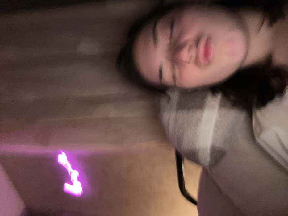 A blurry, low-lit image shows a person lying down, seemingly asleep or resting, with eyes closed and a relaxed facial expression. The background appears to be a wall with some soft, purple light, possibly from a neon sign or similar light source. A blanket with a checkered pattern is visible near the person's shoulder, suggesting a comfortable or cozy setting. The focus is soft, giving the image a dreamlike quality.