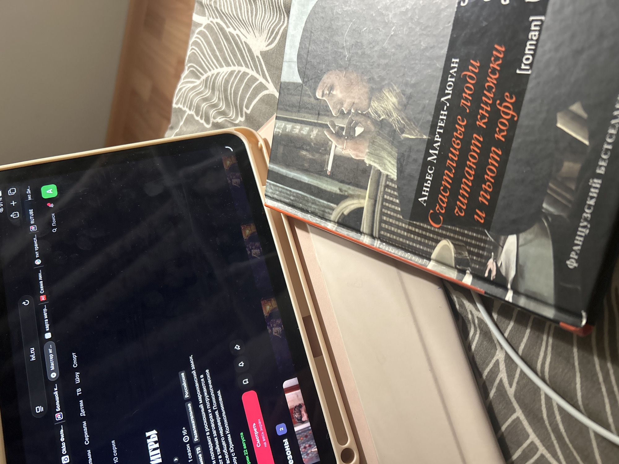 The image shows a close-up view of a tablet displaying a streaming service interface, with a book lying on top of it. The tablet screen shows the website "ivi.ru" and a menu with categories like "Фильмы", "Сериалы", "Детям", "ТВ", "Шоу", and "Спорт". The content displayed on the tablet appears to be a series titled "ИГРЫ" (Games), with details about its season and age rating. There is also a button to "Смотреть на весь экран" (Watch in full screen) dated August 22.
The book is titled "Счастливые люди читают книжки и пьют кофе" (Happy people read books and drink coffee) by Аннес Мартен-Люган. The cover features a woman smoking a cigarette.
The setting appears to be a bedroom, with a patterned gray blanket visible in the background. A white charging cable is also present, connecting to the tablet. There is no visible indication that this scene is located in the city of Троица, Russia.