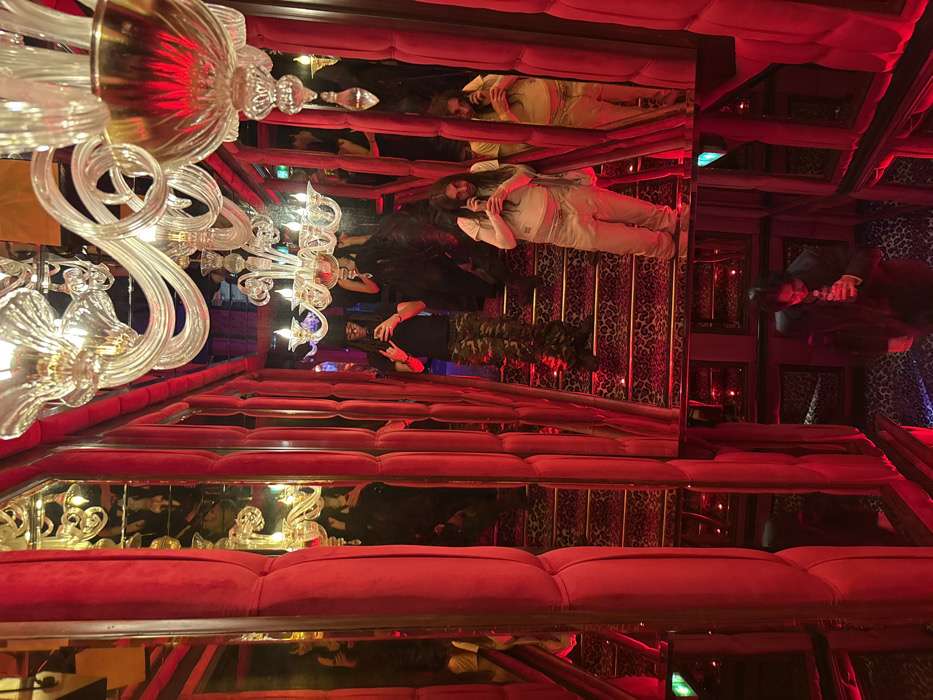 The image depicts the mirrored interior of a staircase or hallway, identified as likely being within the Crazy Horse Paris cabaret, located at 12 Avenue George V, 8th arrondissement, Paris, France. The environment is characterized by predominantly red illumination, extensive mirrored walls creating an infinite reflection effect, and red velvet upholstered panels. Multiple ornate crystal chandeliers with lit bulbs hang from the ceiling, reflecting throughout the space. The stair treads feature a dark, textured pattern, possibly leopard print, accented by gold-colored risers.
Four distinct individuals are visible, alongside numerous reflections of themselves and the environment. In the mid-ground, a person with dark dreadlocks and sunglasses, wearing a black t-shirt and camouflage trousers, stands on a stair tread holding a smartphone in both hands, facing forward as if capturing an image or video. To their right, another person with long dark hair, dressed in a light-colored t-shirt and light baggy trousers, holds a smartphone to their ear. Partially obscured behind the first individual, a third person in dark attire is visible. Towards the bottom of the frame, lower down the reflected staircase, a fourth individual, wearing a dark suit jacket and lighter shirt, holds a small glass or cup to their mouth. The individuals appear engaged with their personal devices or drinks within the visually complex and opulent setting.