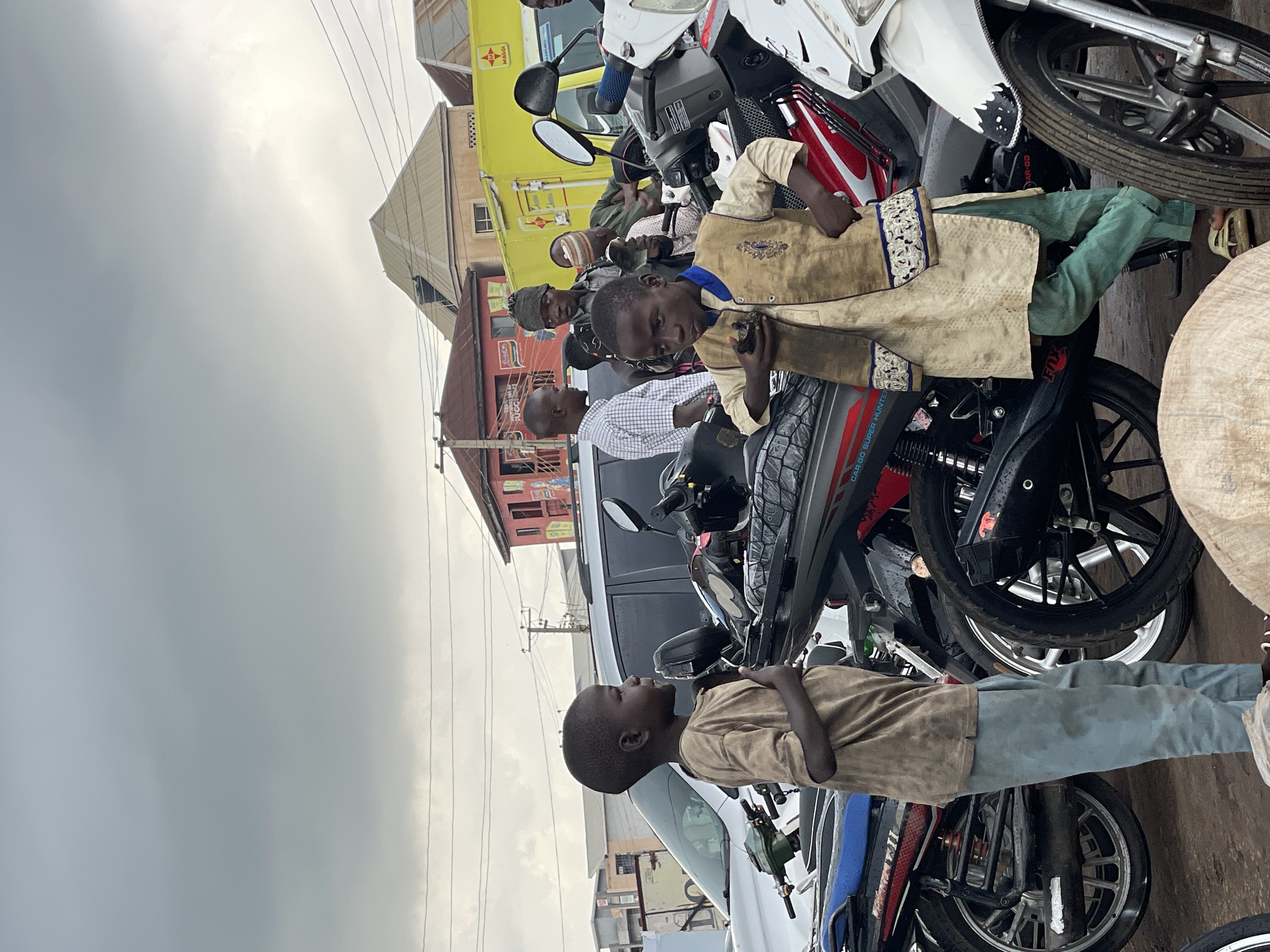 The media file shows a group of people and motorcycles in an outdoor setting, likely a street or market in Jos, Nigeria. The image is oriented vertically, making it appear as if the scene is tilted.
In the foreground, a young boy wearing a tan shirt and light blue pants stands looking to his left. Behind him, a group of people are gathered around several motorcycles. One boy, dressed in a tan garment with blue and white trim, is seated on a motorcycle, looking towards the camera. Other individuals are visible in the background, some wearing hats, and appear to be engaged in conversation or observing the scene.
The objects in the scene are primarily motorcycles of various makes and models, parked closely together. One motorcycle has a prominent red and white design. A white van is also partially visible, parked behind some of the people. Buildings with colorful facades, typical of urban African architecture, are present in the background.
The time of day appears to be daytime, with overcast skies suggesting a cloudy or possibly rainy weather. The overall mood seems casual, with people interacting in a communal space.
Notable details include the detailed embroidery on the tan garment worn by the seated boy. Faint text can be seen on some of the motorcycles, such as "CAP GO SUPER HUNTER" on one. The presence of multiple motorcycles and the general street scene suggest a busy commercial or transportation hub.