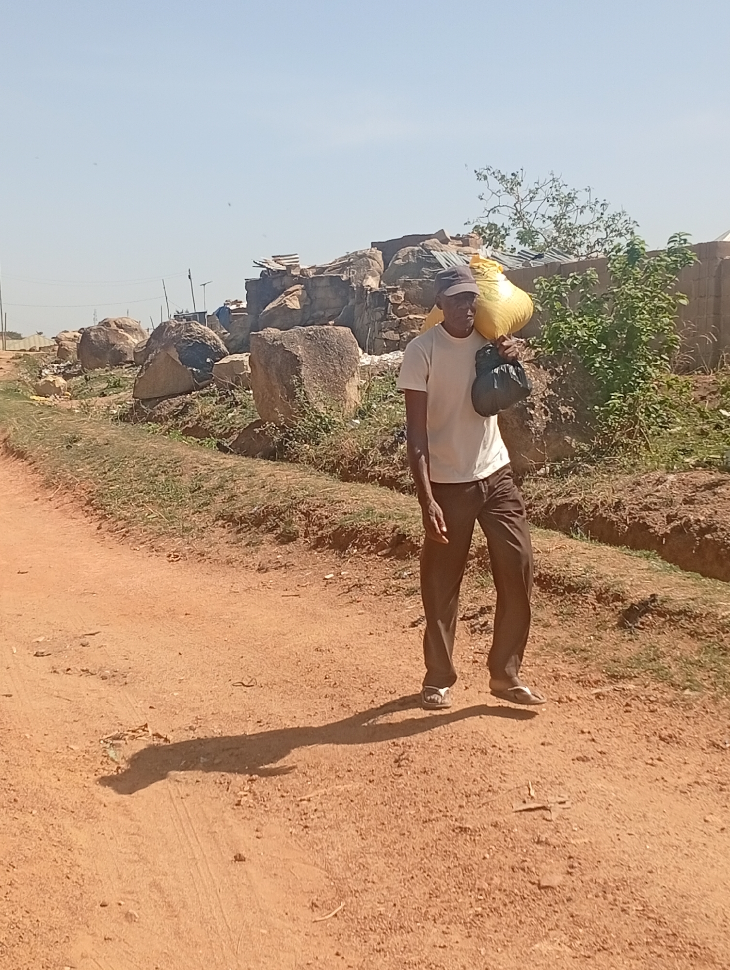In Barakin Akawo, Nigeria, a man wearing a light-colored T-shirt, dark trousers, and a baseball cap is walking on a dirt road. He carries a large yellow sack over his left shoulder and a black bag in his left hand. The man is walking away from a pile of rocks and debris near the roadside. Vegetation and a partially constructed or unfinished brick wall are visible to the right. The sky is clear and light blue. The lighting indicates a bright, sunny day. The road is dry and dusty, with the man's shadow cast to his left. He is wearing sandals.