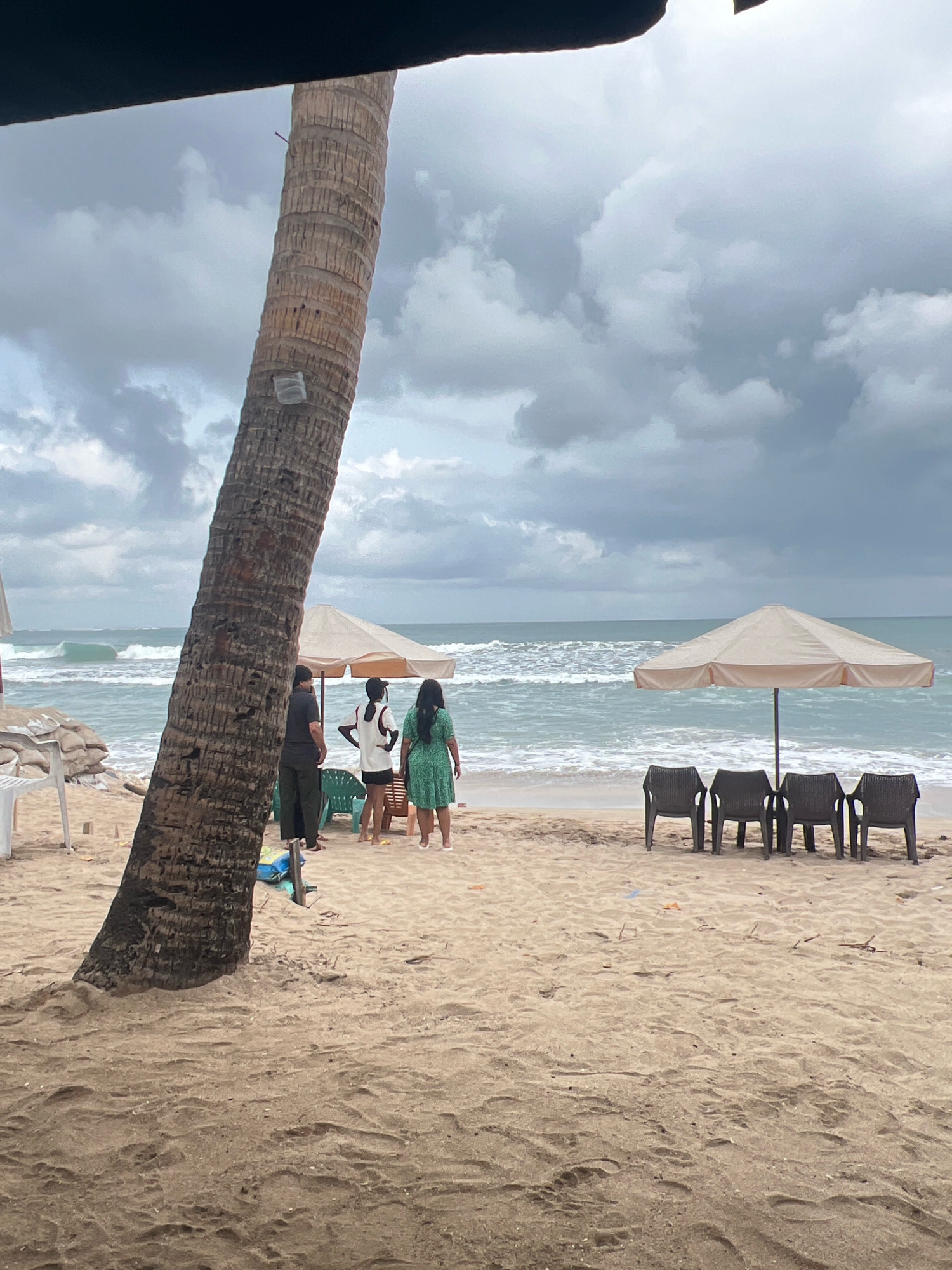 The image depicts a beach scene on an overcast day. A large palm tree trunk is prominently positioned in the foreground, leaning to the left. To the right of the palm tree, several people are gathered on the sand. Three individuals stand together near a beige umbrella supported by a central pole. One person wears a dark shirt and pants, another a white top and dark shorts, and the third a green dress. Further to the right, four dark plastic chairs are arranged in a row facing the ocean, with another umbrella partially visible behind them. The ocean, with moderate waves, stretches to the horizon under a cloudy sky. The sand on the beach shows footprints and scattered debris. To the far left, a white plastic chair and sandbags are visible. The location is Kuta, Indonesia.