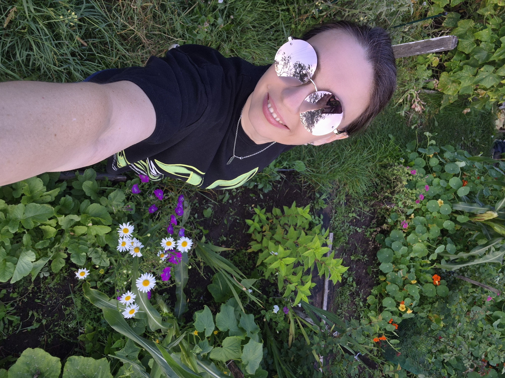 The image shows a woman wearing round, rose-tinted sunglasses and a black t-shirt with a neon green graphic. She is smiling and taking a selfie from a high angle, with her arm extending into the lower left corner of the frame. She is outdoors in a garden setting, with lush green foliage and flowers visible around her. The garden appears to be part of a residential area. The sky is not visible in the frame. The lighting suggests it is daytime, likely late afternoon or early evening. The overall impression is that of a casual outdoor moment. This scene is consistent with a garden located in the city of Chaikovsky, Russia, where such residential gardens are common.