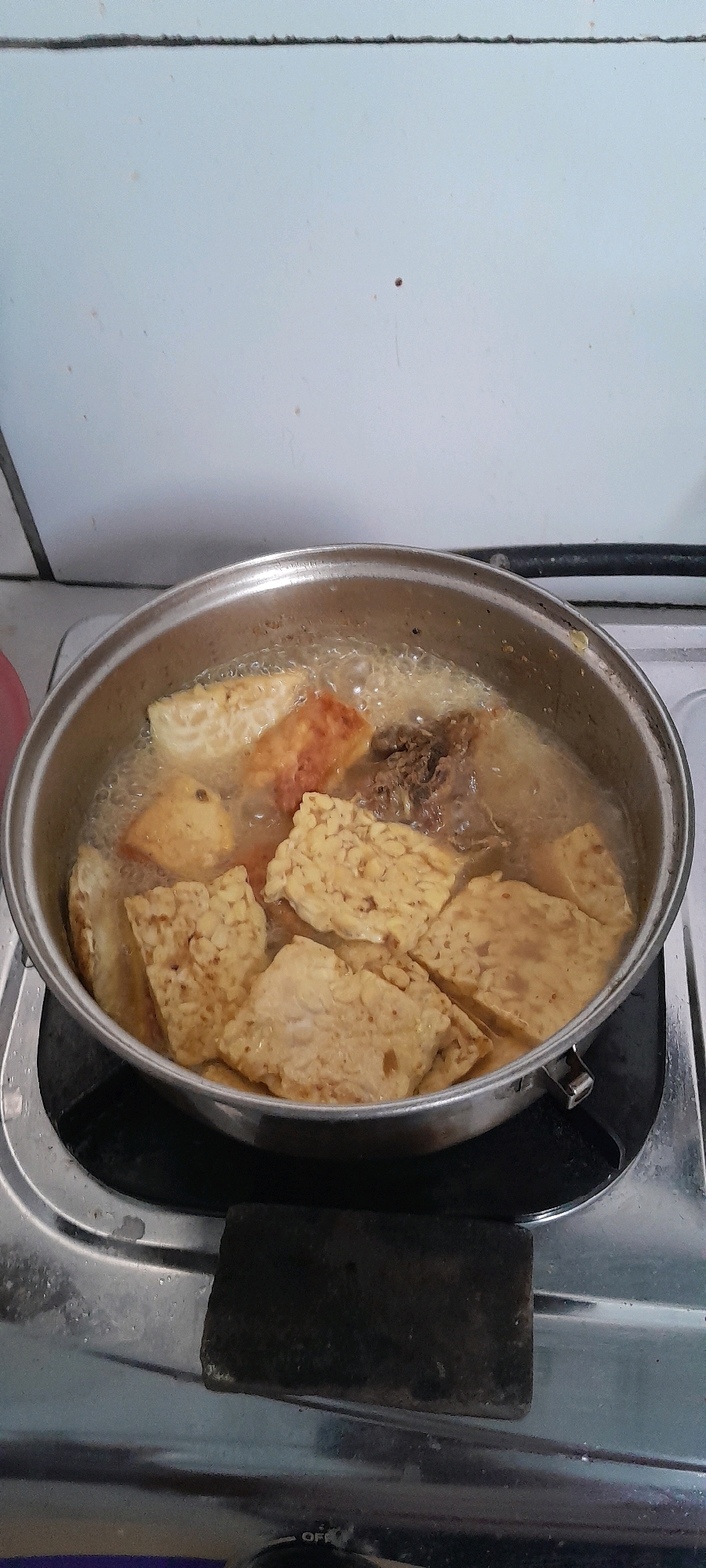 An overhead, slightly angled medium shot shows a stainless steel pot filled with bubbling oil and pieces of fried food, likely tempeh and possibly some meat, being cooked on a gas stove. The pot is situated on a silver stove top. The background consists of a light-colored wall with some faint smudges. The image captures a domestic scene in Bendungan, Indonesia, focusing on the cooking process.