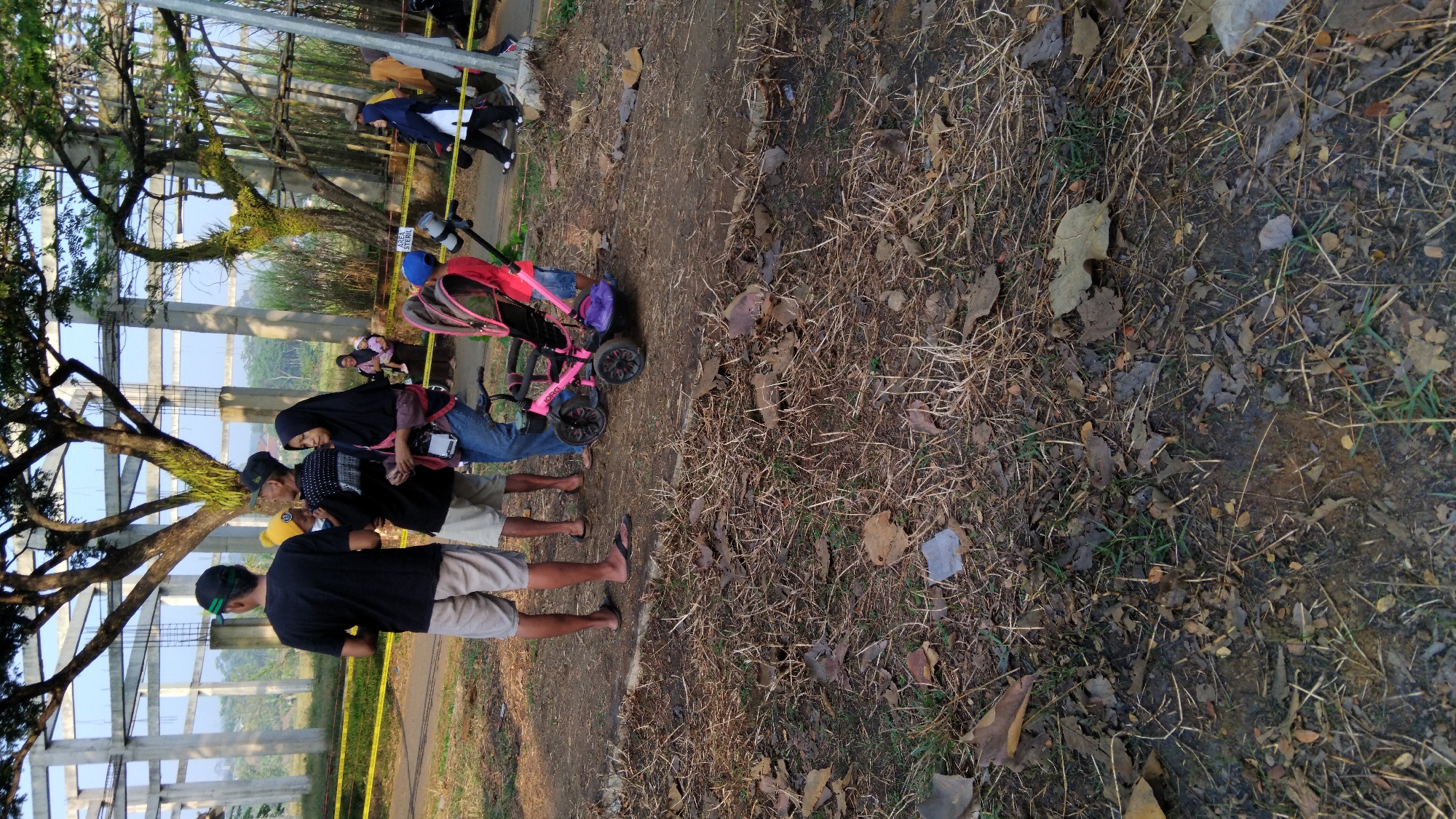 The image depicts an outdoor scene, possibly in the city of Posong, Indonesia. In the foreground, dry grass and fallen leaves cover the ground. A pink stroller with purple accents is prominently placed in the middle ground. Several individuals are visible, some standing near the stroller. To the left, two men and a woman wearing a hijab are conversing. The man on the far left wears a black t-shirt and light-colored shorts, with a green cap. The man next to him wears a black t-shirt and shorts. The woman is in a black hijab and blue jeans. Further back, a group of people, including a woman holding a child, are seen near a yellow barrier tape. In the background, a large, unfinished concrete structure with pillars and beams dominates the scene, suggesting a construction site or a large outdoor event venue. Trees with spreading branches are visible in front of the structure, and more foliage lines the right side of the image. The sky appears clear, indicating daylight.