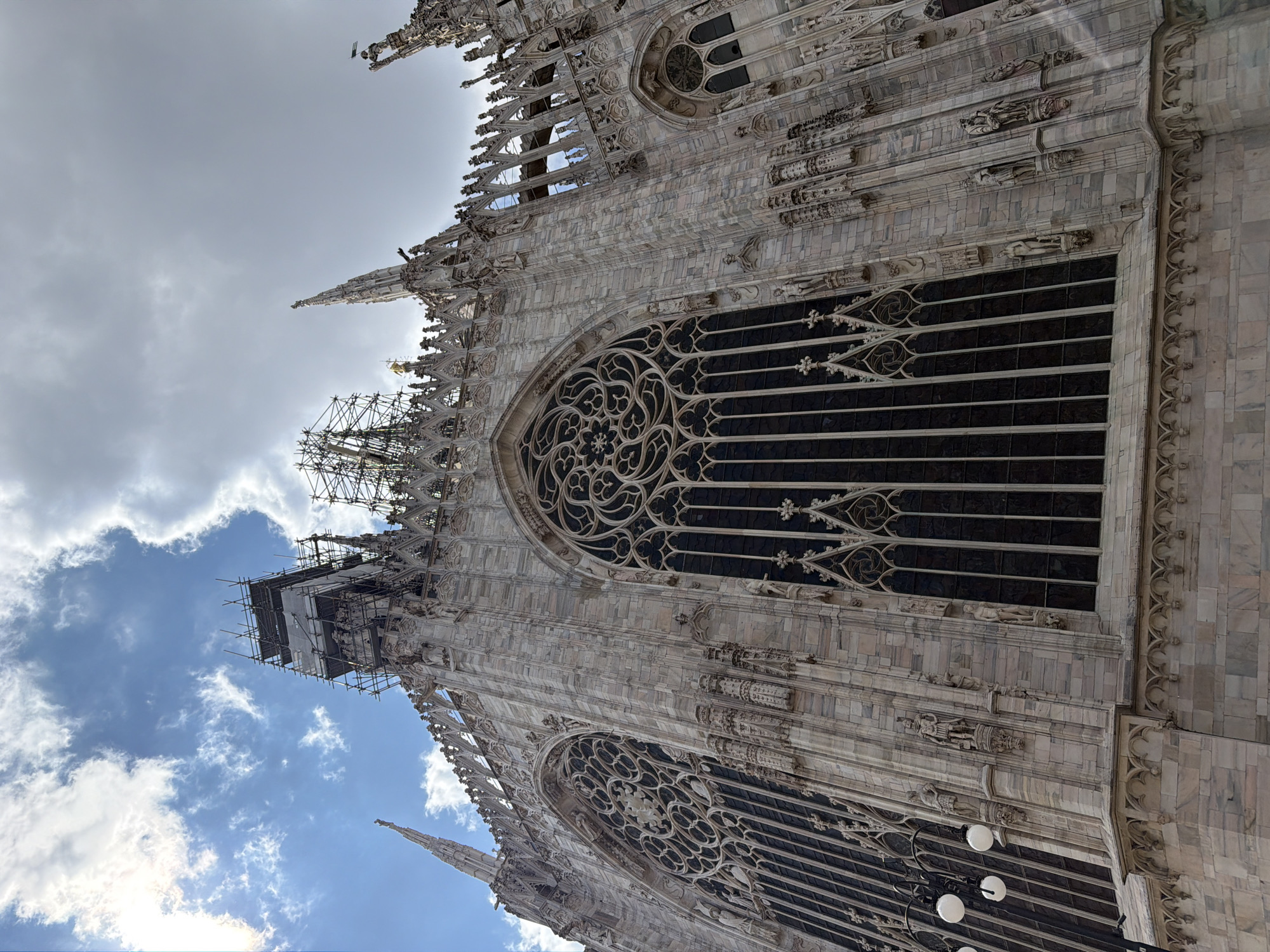 A striking low-angle photograph captures a section of the ornate Gothic facade of the Milan Cathedral (Duomo di Milano) on a partially cloudy day. The immense structure, constructed from white marble, dominates the frame, showcasing its intricate details against a sky that transitions from overcast grey to patches of bright blue with scattered clouds.
The cathedral's architecture is characterized by numerous spires, pinnacles, and buttresses, adorned with countless sculpted figures and decorative elements. Large arched windows, including distinct rose windows with elaborate stone tracery, punctuate the facade. The glass within these windows appears dark, contrasting with the pale marble.
Scaffolding is prominently visible on several sections of the cathedral, particularly towards the upper left, indicating ongoing restoration or maintenance work on the historical edifice. The perspective emphasizes the cathedral's grandeur and towering height. No people or visible text are present in the image.