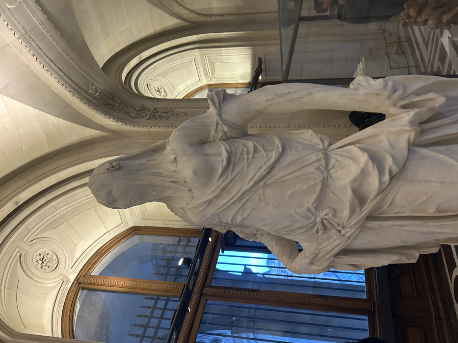 A marble sculpture of a draped figure stands near a large arched window with dark wood framing and metal bars. The window reflects a building across from it. The statue is positioned beneath an ornate, curved ceiling with decorative molding and recessed lighting. The sculpture seems to be displayed behind a glass barrier. The building’s architectural style is elaborate and classical.
