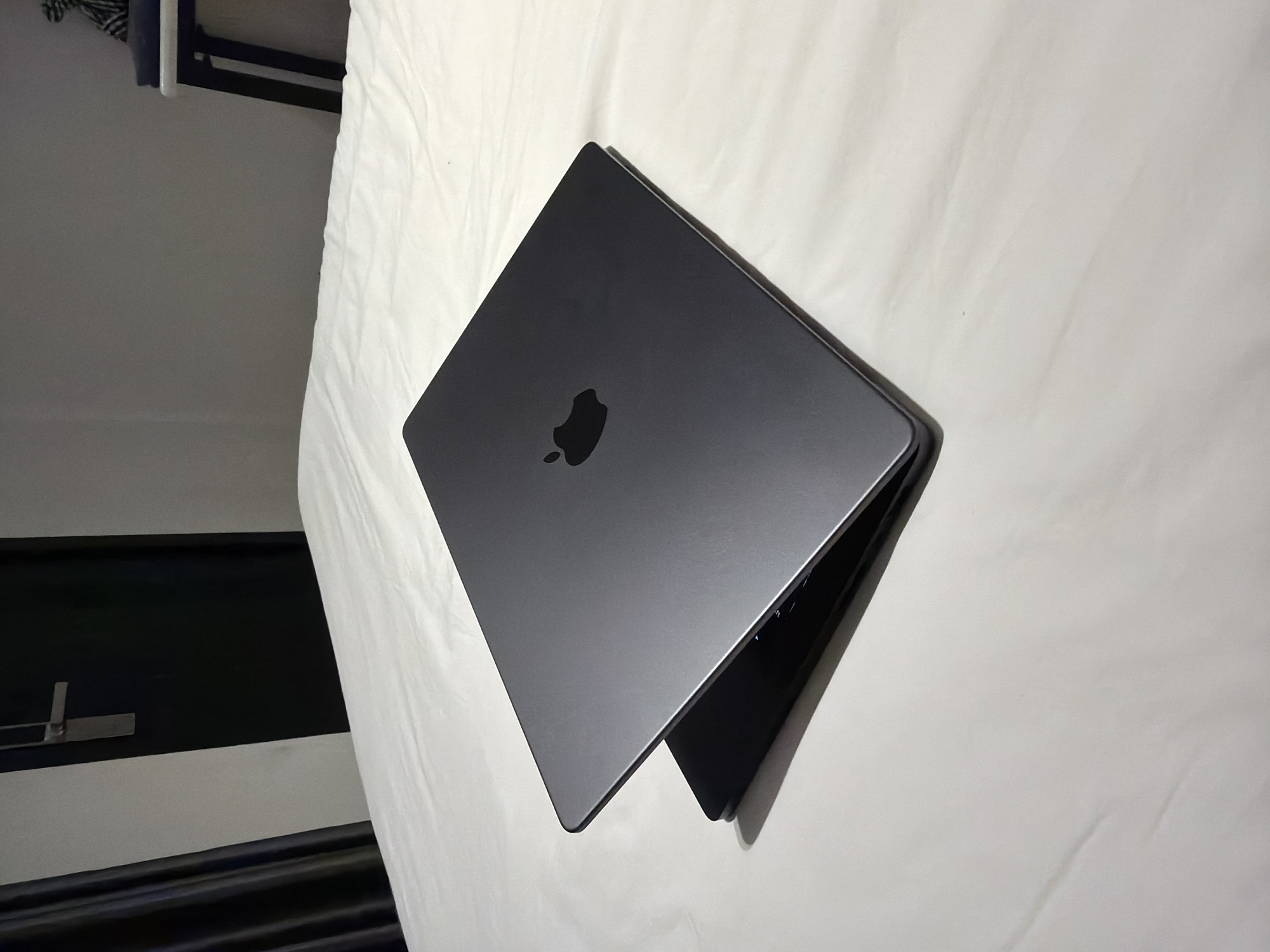 A dark grey MacBook laptop is open and angled on a white bed. The laptop is positioned diagonally with the screen slightly ajar from the keyboard. The Apple logo is visible on the lid of the laptop. The background consists of a dark door with a silver handle, a dark curtain on the left, and a white table with objects on it on the right. The scene is lit with ambient light, casting shadows on the bed.