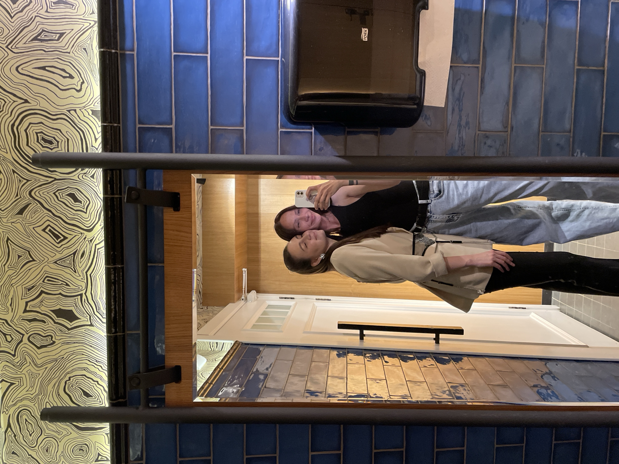 The image depicts two young women taking a mirror selfie in what appears to be a restroom. The woman on the left is wearing a tan jacket over a white top and black pants, with her hair pulled back. The woman on the right is wearing a black tank top and light blue jeans. They are both smiling and looking at the phone. The background features blue tiled walls, a wooden door, and patterned wallpaper. The scene is located in the city of Izhevsk, Russia.