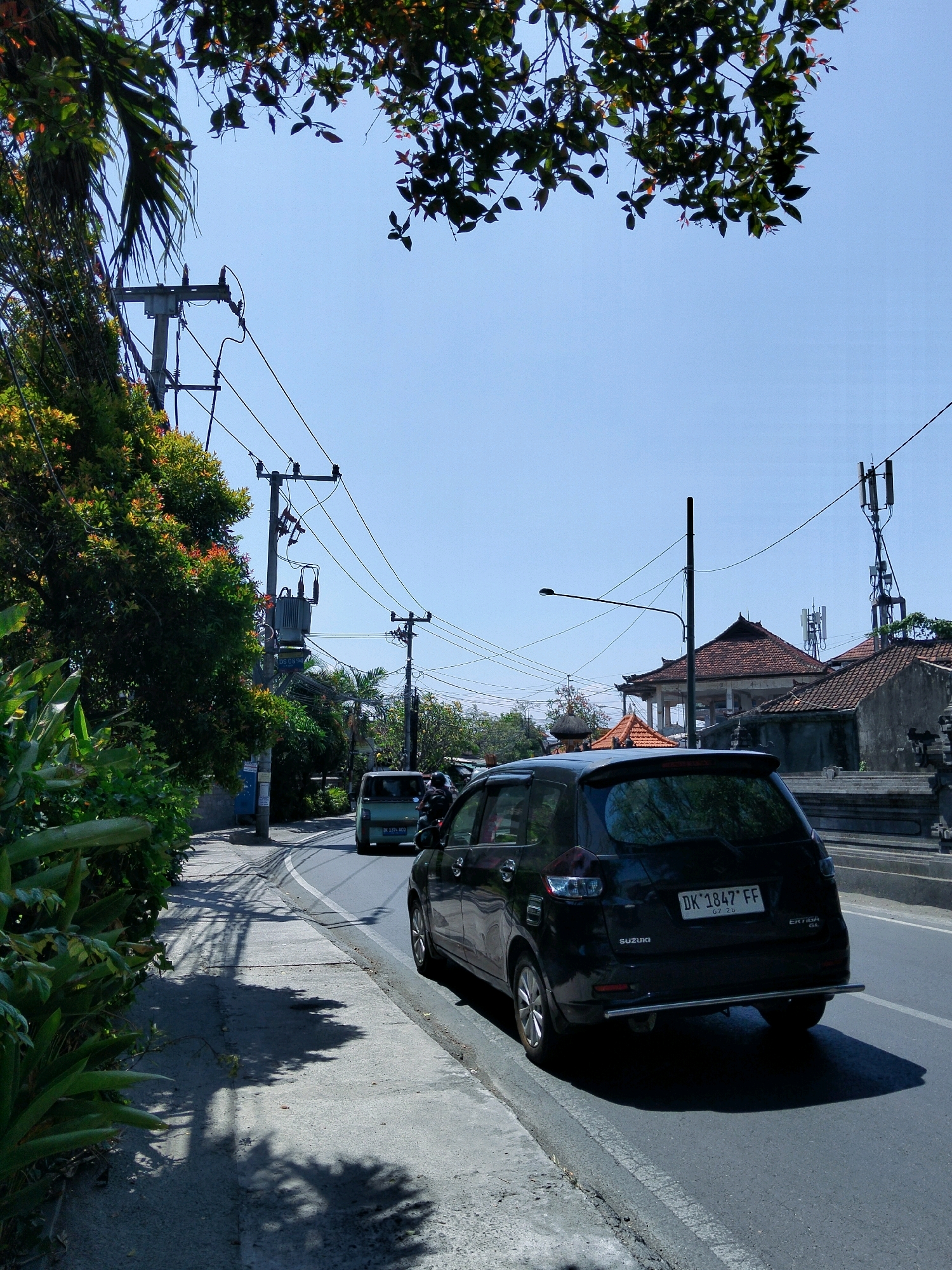 A black Suzuki Ertiga GL with license plate DK 1847 FF is parked on the right side of a two-lane road in Denpasar, Indonesia. The car is viewed from the rear, with its taillights and a chrome bumper visible. A green car is in front of it, also moving in the same direction. The road is paved, with a concrete sidewalk on the left. Lush green vegetation lines the sidewalk, with the shadows of palm trees cast on the ground. Several utility poles with electrical wires crisscross the scene. In the background, traditional Balinese-style buildings with red tile roofs are visible. The sky is clear and blue.