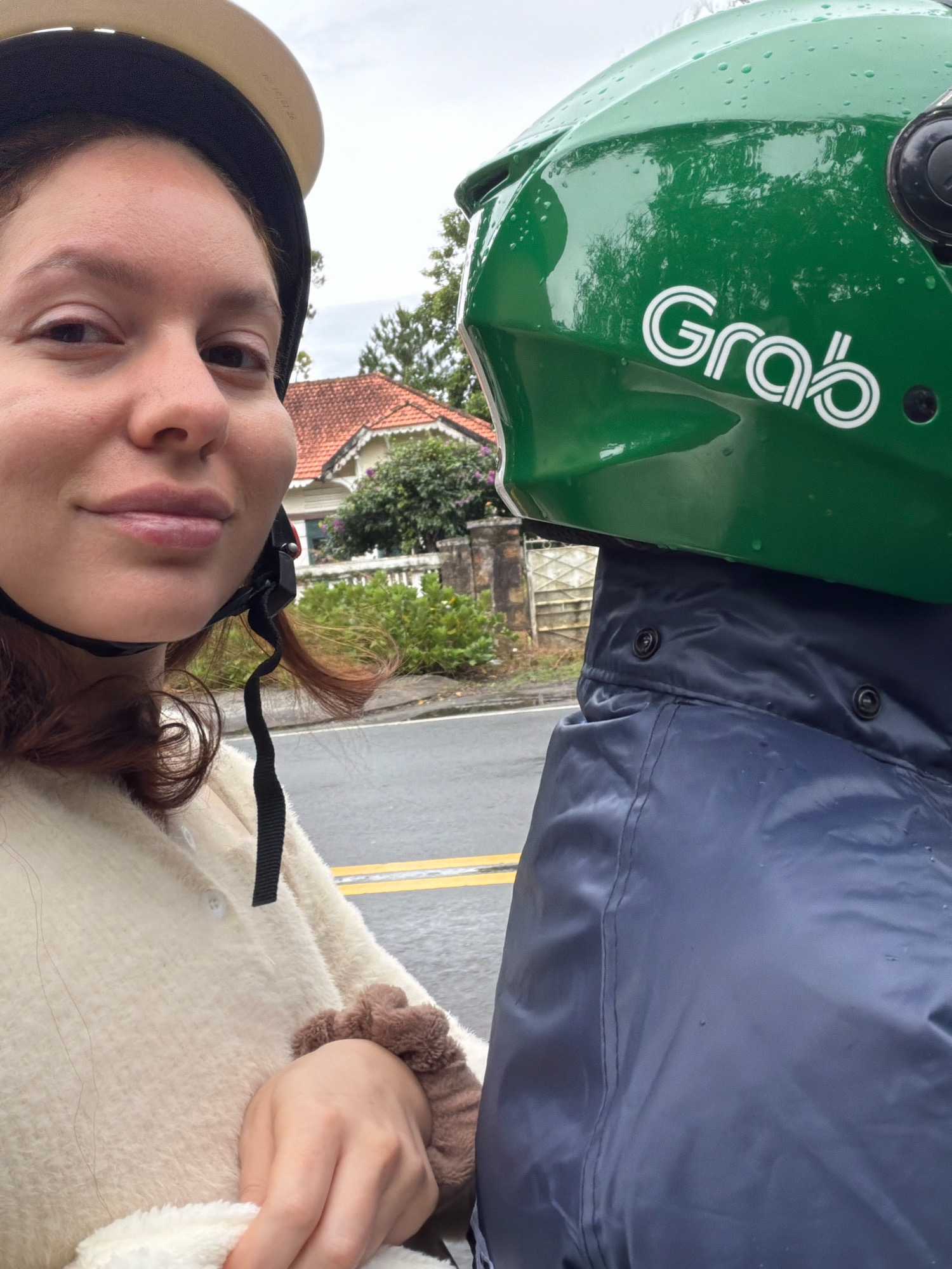 In Da Lat, Vietnam, a young woman wearing a cream-colored fuzzy sweater and a helmet poses for a selfie on a motorcycle. The helmet is beige with a black strap. Beside her is the distinctive green helmet of a Grab driver, with the "Grab" logo clearly visible. The driver, wearing a dark blue rain jacket, is partially visible behind the woman. The background reveals a street with yellow lane markings, suggesting a rainy day, with a house featuring a terracotta roof and lush green foliage. The overall scene depicts a common mode of transport in Da Lat, highlighting the presence of the Grab ride-sharing service.
