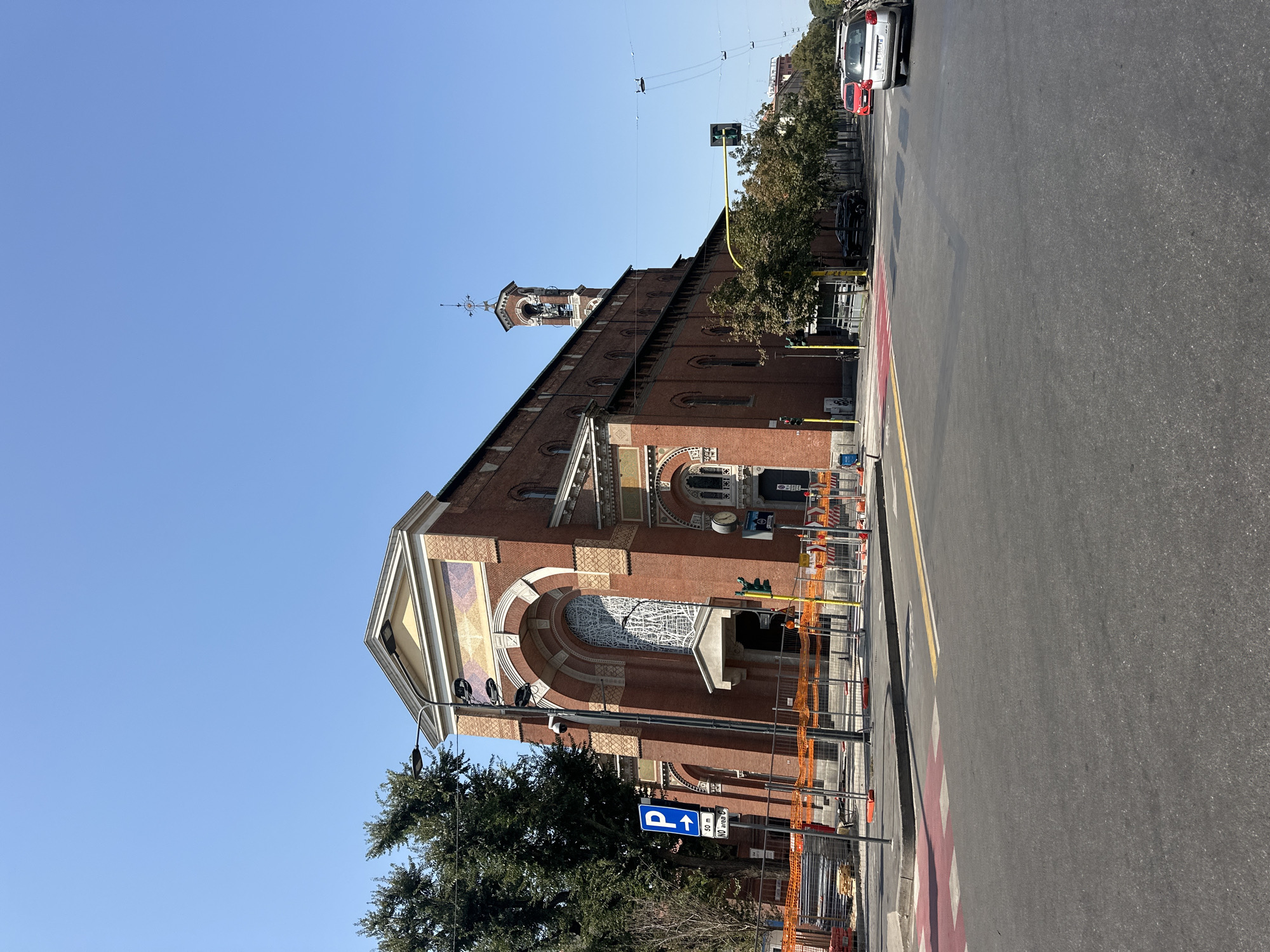 The image captures a wide-angle, street-level view of the Church of San Maurizio al Monastero Maggiore in Milan, Italy. The facade of the red brick church is prominently featured on the left side of the frame, characterized by its large arched window with stained glass and decorative mosaic panels above. A bell tower is visible extending from the right side of the church. In the foreground, the scene depicts a street intersection with a pedestrian crossing marked with red and white stripes. Orange construction barriers line the sidewalk in front of the church, suggesting ongoing work. Traffic lights and street lamps are present, and several cars are visible on the road, including a silver station wagon in the right lane. Trees line the street, and the sky is a clear, bright blue. The overall impression is of a sunny day in an urban environment, focusing on the architectural details of the church and the surrounding streetscape.