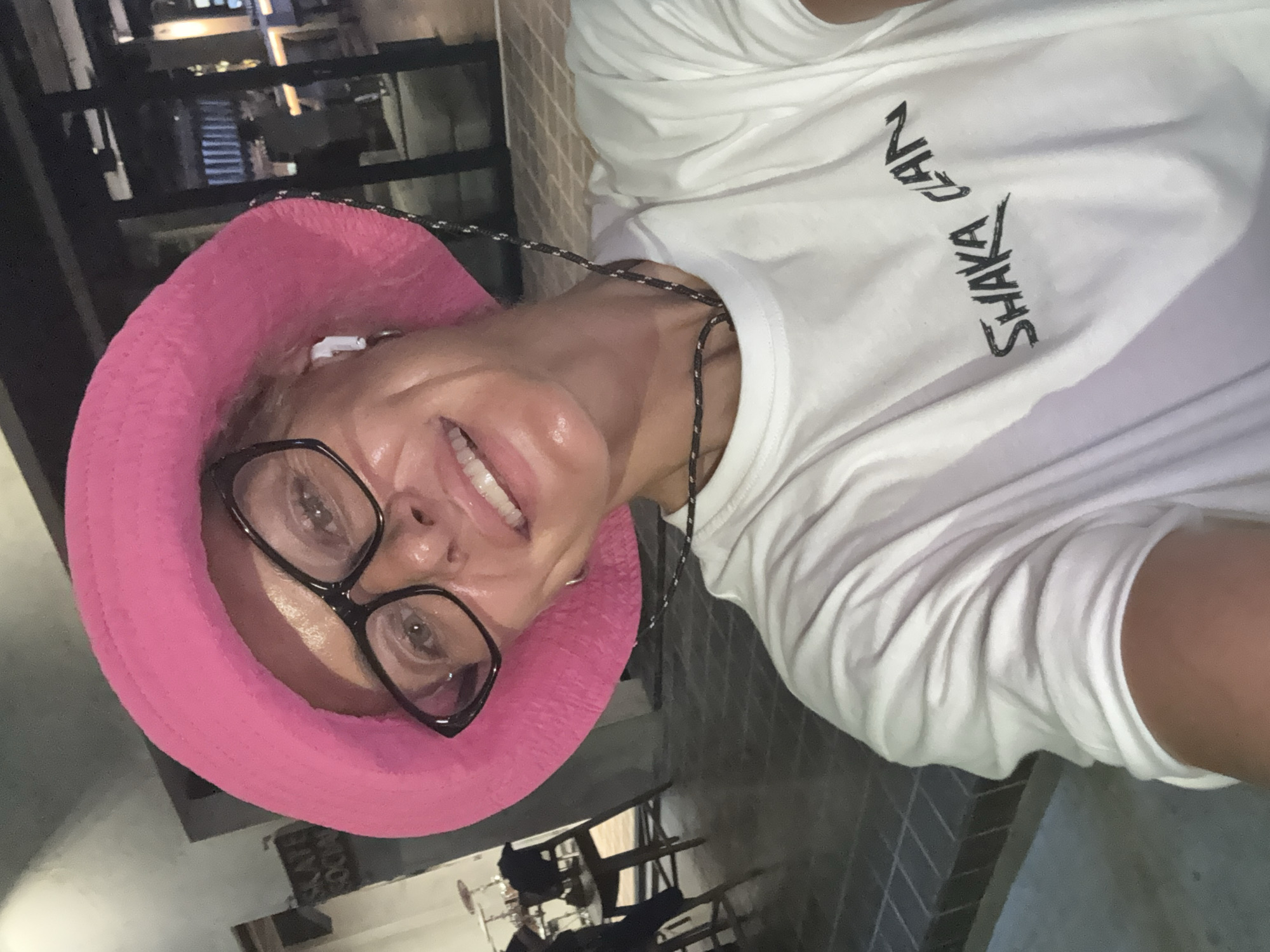 A woman is seen in an indoor setting, wearing a bright pink hat and glasses, with a white t-shirt that has "SHAKA CAN" written on it. The location has a dark and blurry background with what appears to be outdoor lights visible through a glass enclosure. The woman is positioned such that the photograph is taken from a high angle.
