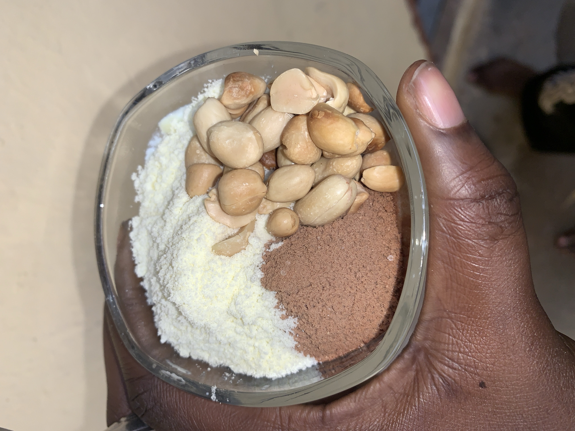 A close-up, top-down shot shows a clear glass bowl held in a dark-skinned hand. The bowl contains three distinct sections: a pile of blanched peanuts, a mound of white powder (possibly milk powder), and a pile of brown powder (likely cocoa or chocolate powder). The photograph's location and city within Nigeria, Rafin Par, are unspecified, and no further information about people, interactions, or the environment is discernible.
