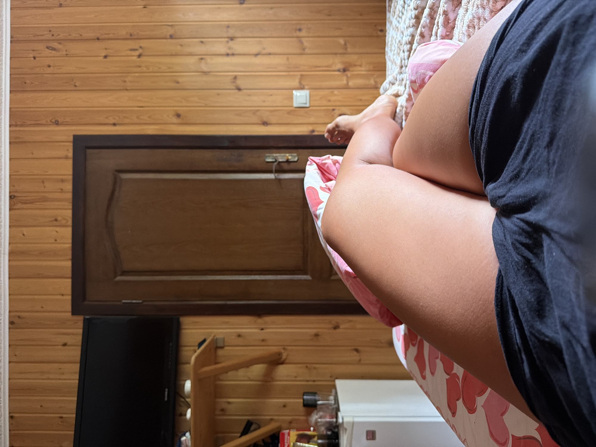 The image depicts a close-up view of a person's legs, one in a black t-shirt and the other partially covered by a pink and white heart-patterned blanket. The person appears to be lying down or sitting in a room with wooden paneled walls. A dark wooden cabinet or door is visible on the wall behind the person, with a small, square light switch above it. To the left, a black flat-screen television is mounted on the wall, and a wooden stool or chair is situated below it. In the lower right corner, a white appliance, possibly a washing machine or dryer, is partially visible, along with some items on a shelf. The scene suggests an indoor setting, likely a bedroom or living area, with a casual and relaxed atmosphere. The lighting indicates it is daytime. There is no visible text in the image.