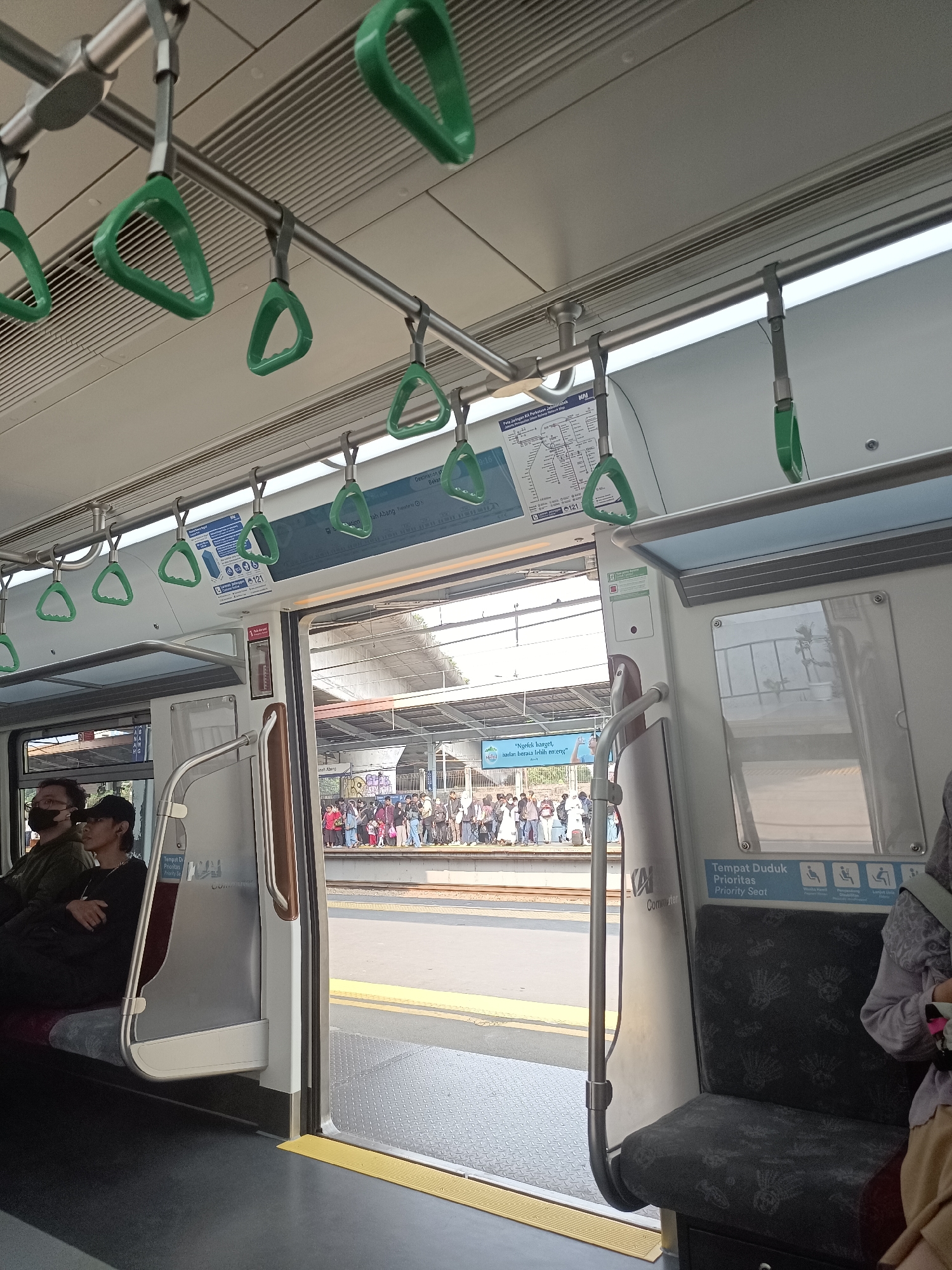 The image depicts the interior of a commuter train at Sudirman Station in Central Jakarta, Indonesia. The perspective is from inside the train, looking outwards towards the station platform and a crowd of people waiting. Green hand straps are visible hanging from the ceiling, a common feature in public transport for passengers to hold onto.
Inside the train, two individuals are seated on the left side. The person in the foreground is wearing a dark shirt and a black face mask, with glasses. Next to them, another person is wearing a black baseball cap and a dark shirt. On the right side of the train, a priority seat is visible, with a sign indicating "Tempat Duduk Prioritas" (Priority Seat). A person wearing a patterned purple top and a beige skirt is partially visible sitting in this area.
Through the open doors of the train, the station platform is clearly visible. A substantial number of people are standing on the platform, appearing to be waiting for the train or disembarking. The platform has yellow and grey markings. The station architecture appears modern, with a covered roof supported by pillars. Graffiti is visible on a structure behind the crowd on the platform. The lighting suggests it is daytime, with sunlight casting shadows. The overall environment is that of a busy urban public transportation hub.
