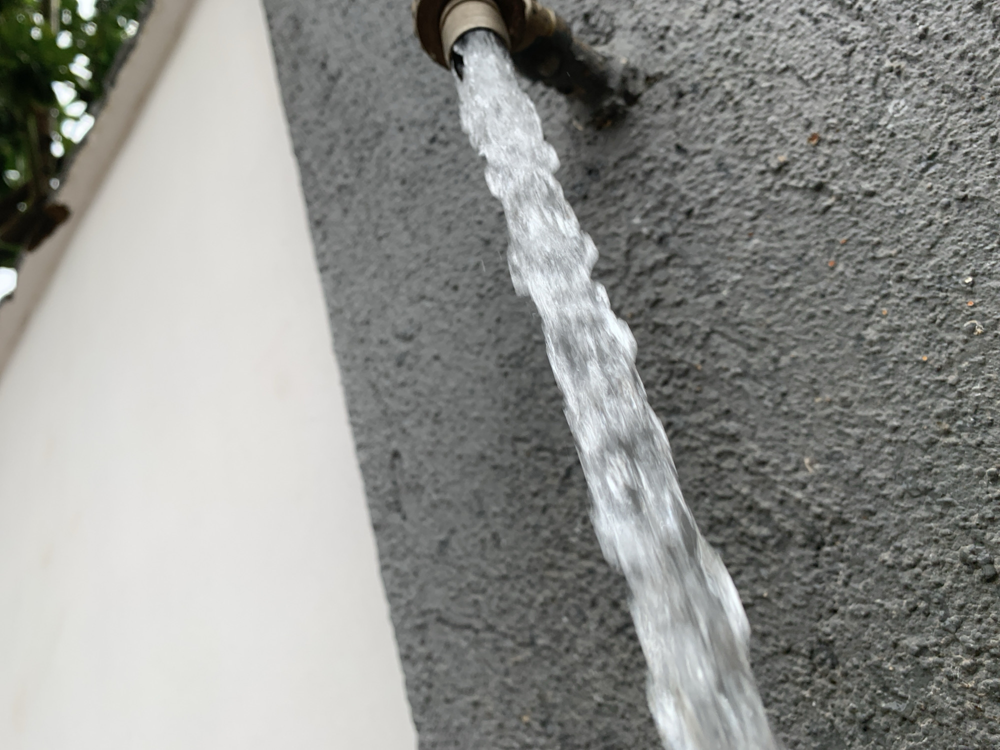 A close-up, low-angle shot shows water streaming from a brass faucet attached to a rough, textured gray wall. The water forms a coherent, bubbly stream with visible eddies and ripples. To the left, a portion of a smooth, off-white wall and some blurred green foliage are visible, suggesting an outdoor setting. The faucet itself is partially visible, as is a section of pipe extending into the wall. The scene appears to be in Gura Top, Nigeria, an area known for its varied terrain and proximity to natural water sources, possibly indicating a residential or public outdoor faucet.