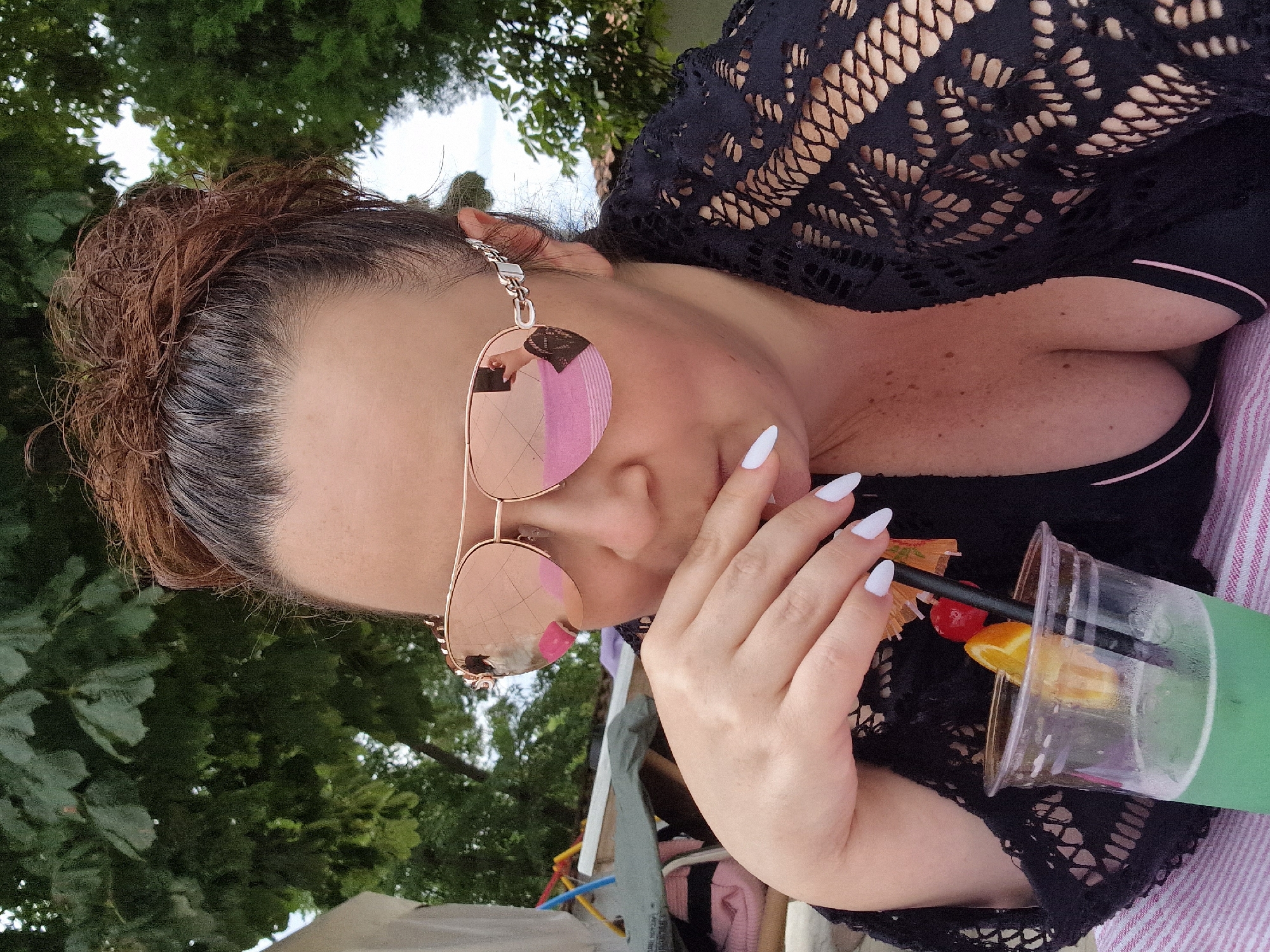 A close-up, medium-shot, over-the-shoulder photograph captures a woman in Kazanlak, Bulgaria, enjoying a drink. She is wearing sunglasses with rose-tinted lenses and a black lace cover-up over a black bikini top. Her hand, with white-tipped nails, is raised to her mouth as she drinks from a clear plastic cup containing a green beverage, a slice of orange, a cherry, and a decorative umbrella. The background reveals lush green trees and the blurry outlines of other people and beach paraphernalia.