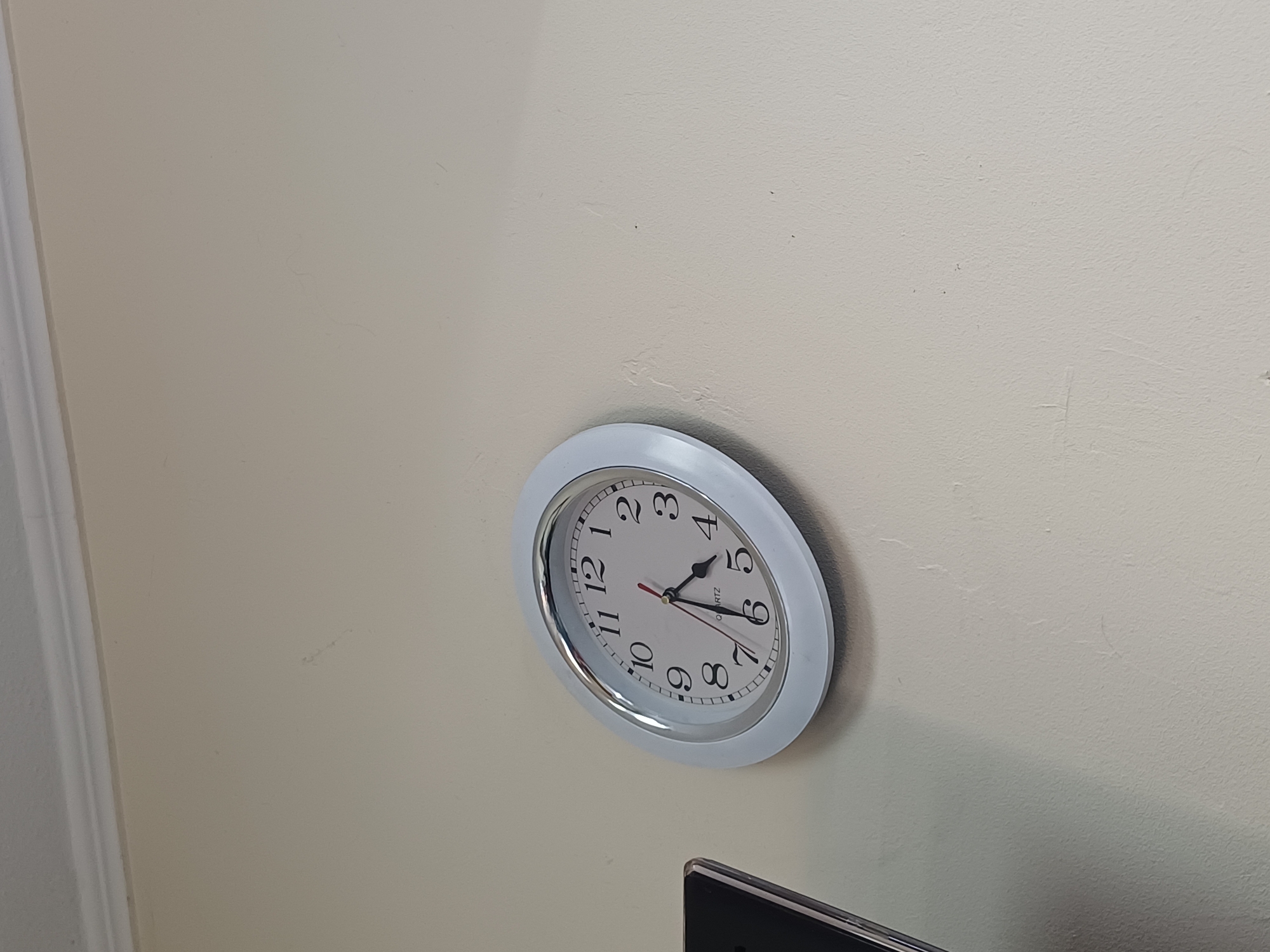 A white, oval-shaped wall clock with a silver bezel is mounted on a cream-colored wall. The clock face is white with black Arabic numerals and black hour and minute hands. The hour hand is pointing to the 4 and the minute hand is pointing to the 6. The clock is slightly tilted to the right. To the left of the clock, a portion of a black electronic device with a screen is visible. The image appears to be taken indoors, in an office or residential setting. There is no indication of the city of Kyiv, Ukraine, or any other specific location.