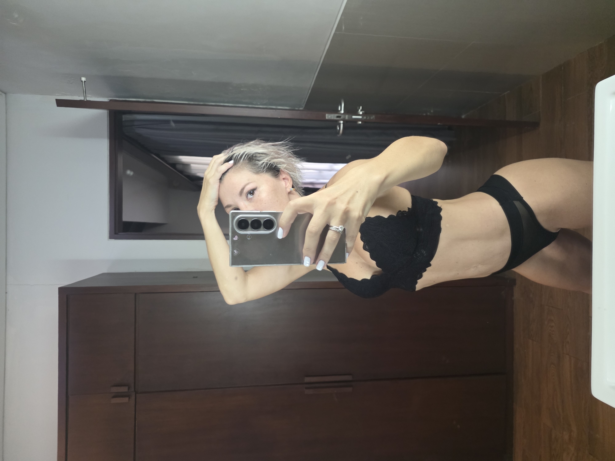 A person is standing in a bathroom and taking a selfie in a mirror. The person is wearing a black lace bra and black underwear. They have short, blonde hair and are holding their phone up to take the picture. The bathroom has wooden floors, a dark brown wardrobe, and a grey tiled wall. A white sink is visible in the foreground. The location is identified as North Kuta, Indonesia.