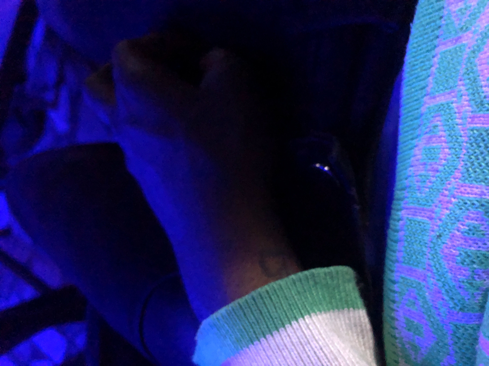 The image displays a close-up, angled view of an individual's arm and upper legs/lap area, illuminated predominantly by blue ambient light. The scene is located in Unknown, Unknown.
A left forearm is visible on the left side, wearing a long-sleeved garment with a cuff featuring horizontal stripes of white and green knit. A faint, dark, circular or heart-shaped tattoo is visible on the inner forearm, extending from beneath the cuff.
The hand rests flat on what appears to be a dark fabric covering the upper thigh or lap. The skin tone of the arm and hand is dark, either naturally or due to the blue lighting conditions.
In the lower-center, partially obscured by the resting hand, a small, reflective object, possibly metallic or glass, displays two distinct points of light.
The lower foreground is occupied by a knitted textile, likely a blanket or garment, displaying a repeating geometric pattern in light blue and purple threads against a turquoise or light green base. These colors appear to fluoresce under the blue illumination. The background is indistinct and very dark.