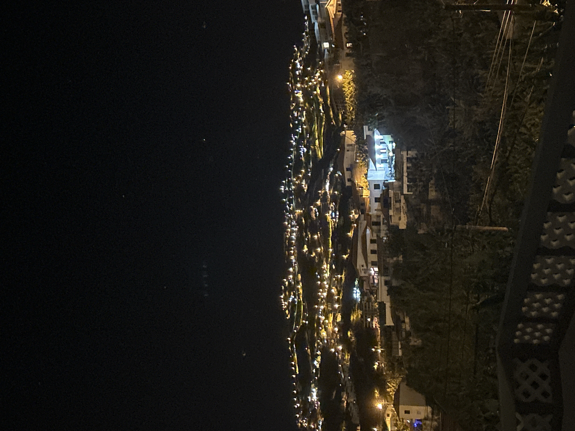 The image shows a nighttime view of Câmara de Lobos, Portugal. The left side of the frame is dominated by the dark expanse of water. The town sprawls across the center of the frame, with numerous lights indicating buildings and streets ascending the hillside. Vegetation is visible intermittently among the buildings. A decorative architectural element partially obscures the view on the right side of the frame, while power lines are visible in the upper right quadrant.