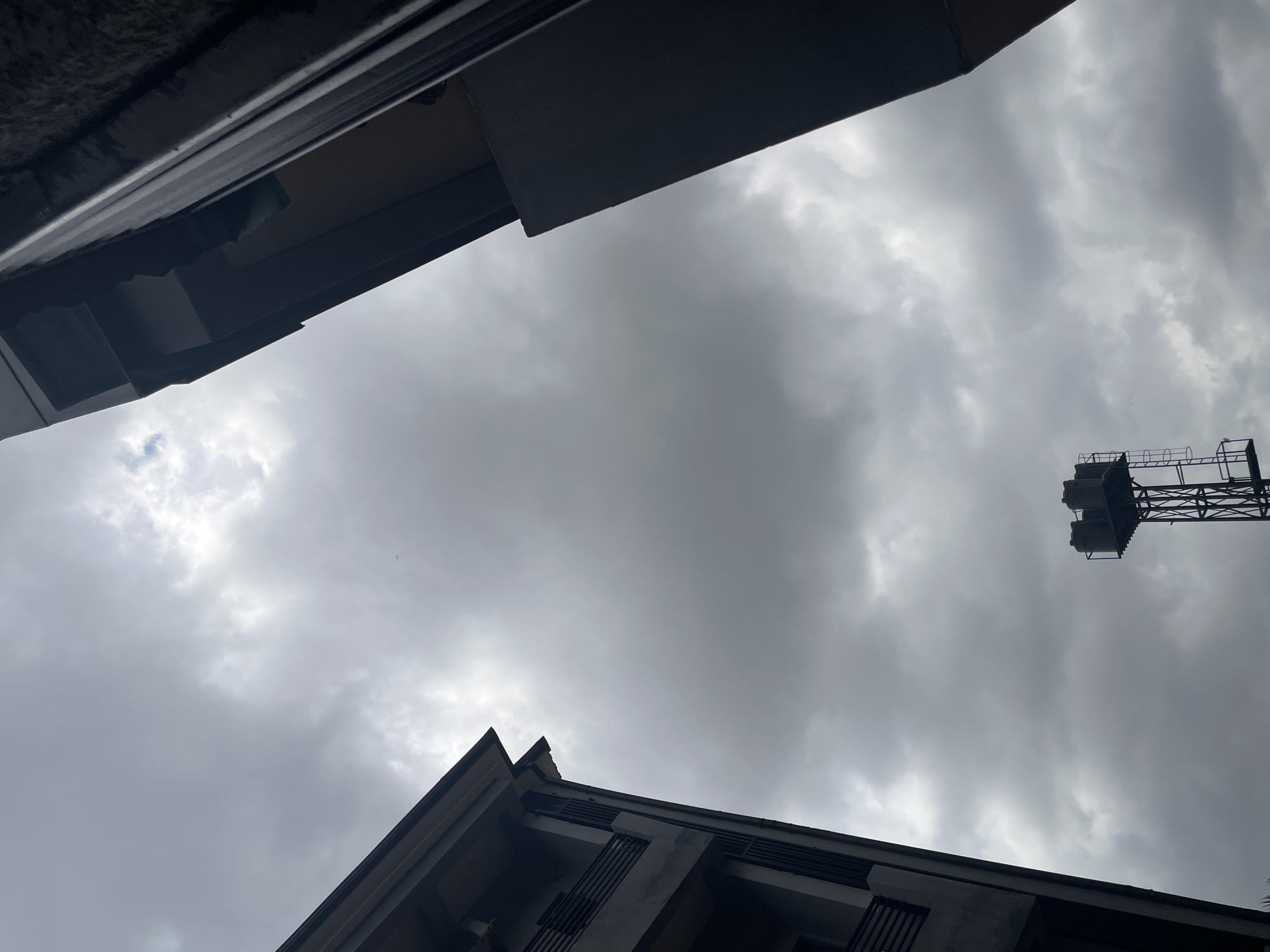 The photograph captures a low-angle view of a cloudy sky above an urban landscape in Abuja, Nigeria. Two buildings frame the sky, one on the left and one on the right, angled inwards. The building on the left is a multi-story structure with visible balconies and dark railings. The building on the right appears to be a more modern structure with a darker, more solid facade. In the center of the lower portion of the image, a water tower with multiple tanks is visible against the cloudy sky. The overall atmosphere is overcast, suggesting a lack of direct sunlight. There are no people or animals visible in the frame. The scene is static and devoid of any overt action.