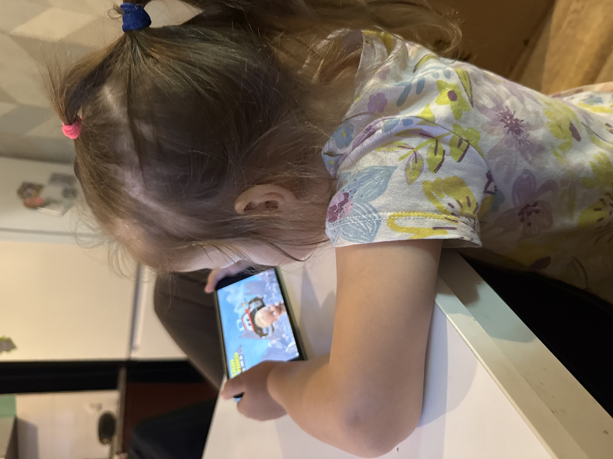 A close-up, side-profile shot shows a young child with brown hair tied into two pigtails, one with a pink hair tie and the other with a blue one. The child wears a light-colored shirt with a floral pattern of purples, yellows, and blues. They are looking down at a smartphone held in their hands, which displays a cartoon image of a character in an aviator outfit. The child's arm is resting on a white surface, possibly a table, and the background is blurry but suggests an indoor setting with hints of furniture and possibly shelves. The lighting appears to be artificial. No specific time of day or weather conditions are discernible. The child's expression is focused on the screen. There is no visible text in the image.