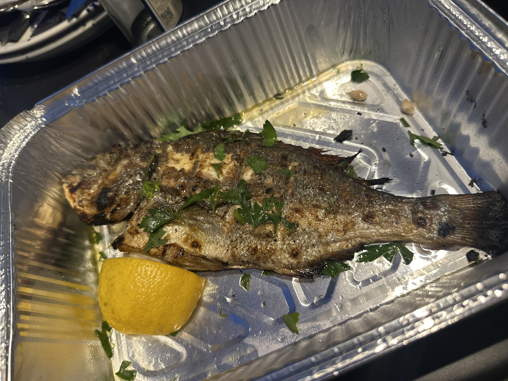 The image shows a whole grilled fish, likely a sea bream or similar species, served in a foil takeout container. The fish is seasoned with herbs and appears to be seasoned with salt and pepper. A wedge of lemon is placed next to the fish. The container is slightly oily, and there are some droplets of liquid visible. The background is dark and out of focus, suggesting the photo was taken in low light conditions. No people are visible in the image. The setting does not provide any specific clues about its location in Leptokarya, Greece, beyond the presence of a takeout container for food.