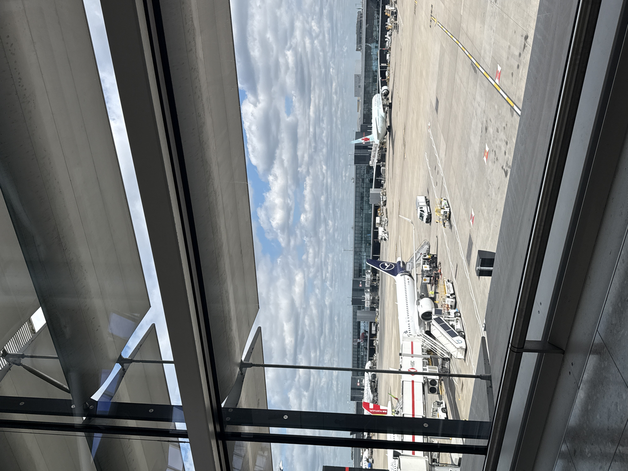 The image depicts a view through a window at what appears to be an airport, possibly located in London, United Kingdom. Several airplanes are visible on the tarmac, with ground service equipment surrounding them. One airplane has the livery of Lufthansa. Another has the livery of Air Canada. The sky is overcast with many clouds. The window through which the scene is viewed has a series of slatted blinds. The overall environment suggests a busy airport terminal setting.