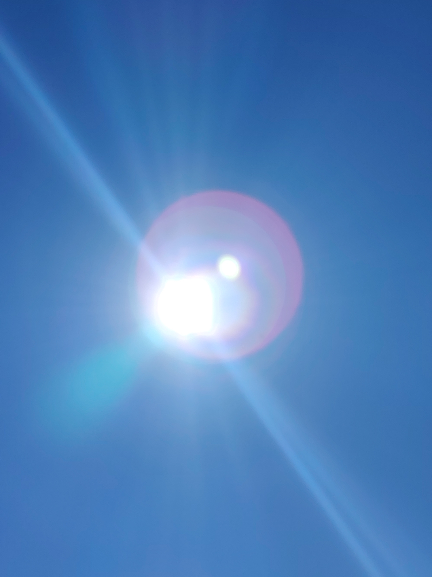 The image shows a bright light source, possibly the sun, in a blue sky. The light has a prominent lens flare and surrounding diffraction effects. There are no identifiable people, subjects, or objects in the image beyond the light source and the sky. The environment appears to be a clear, sunny day. The image does not provide enough detail to confirm a specific location in Noviy, Russia.