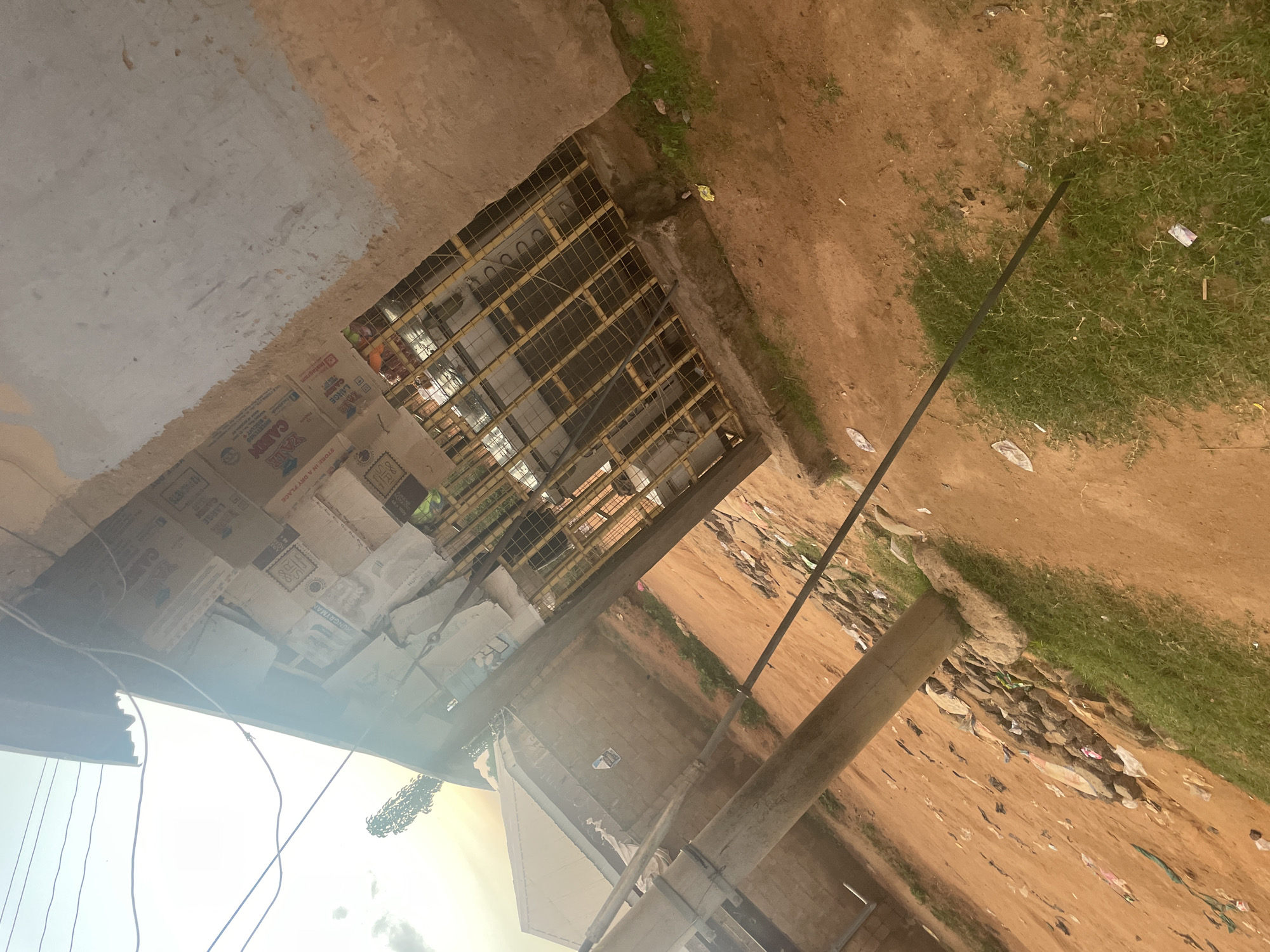 This image depicts a rustic, outdoor market stall, likely situated in the city of Jalingo, Nigeria. The stall is constructed with a corrugated metal roof and a rough, unfinished concrete or plaster wall. The front of the stall is covered by a yellow metal grate, behind which goods are displayed. Some of the merchandise appears to be packaged in cardboard boxes with text that includes "CABIN" and "LARGE." The ground in front of the stall is a reddish-brown dirt, with patches of green grass and scattered debris, including rocks and pieces of plastic. A thick, wooden pole stands in the foreground, partially obscuring the view of the stall. Overhead power lines are visible against a bright sky, suggesting an urban or semi-urban setting. The overall impression is one of a functional, but somewhat neglected, commercial space in an informal economy.