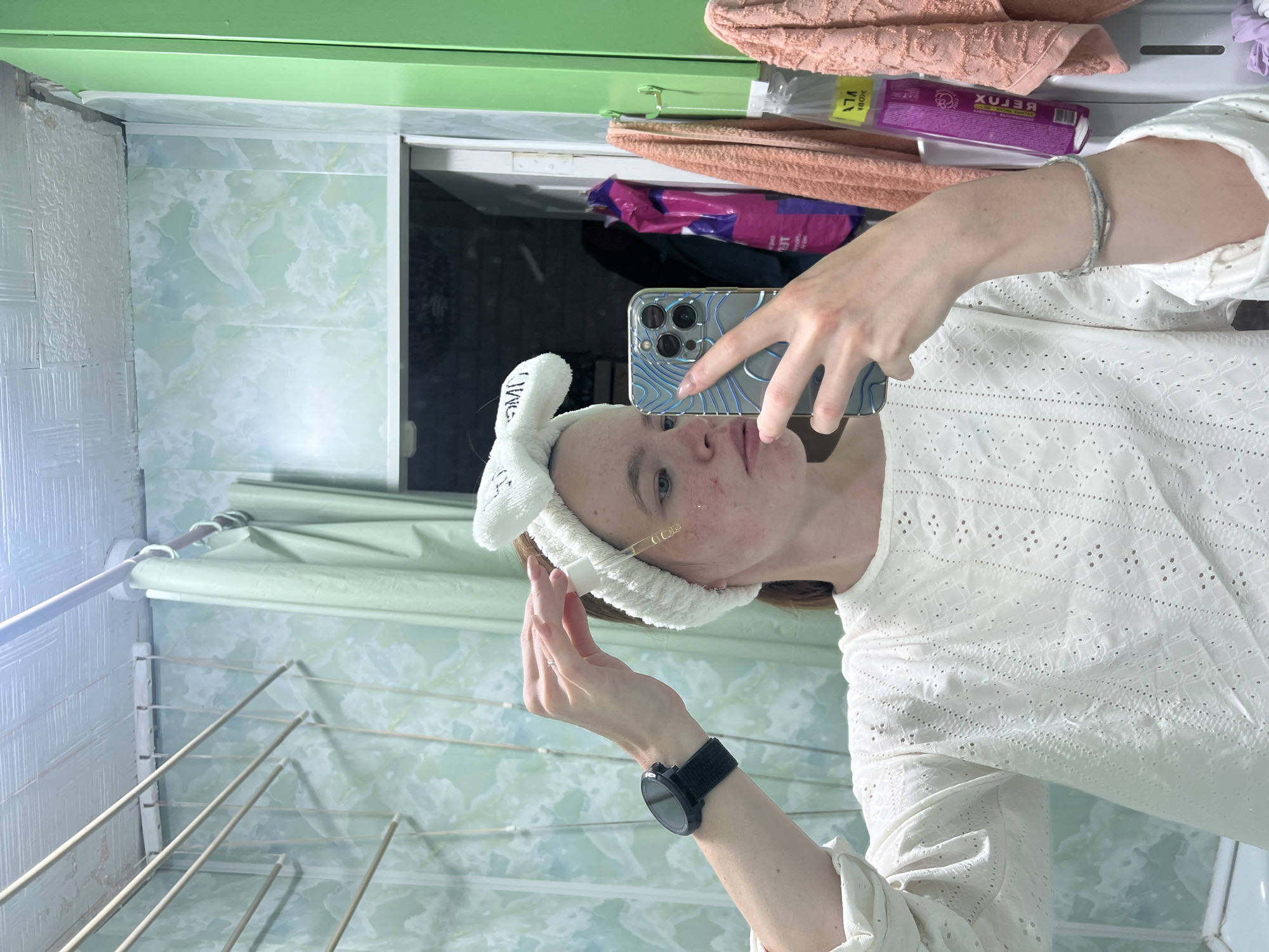 A person with a white bunny ear headband is taking a selfie in a bathroom. The person is wearing a white eyelet blouse and a smartwatch on their left wrist. They are holding an iPhone with a patterned case in their right hand. A gold-colored dropper is near their face.
Behind the person, there is a wall with light green patterned tiles and a drying rack. Above the person's head, there is a shelf with towels and a box of tissues.
The location is Chaikovsky, Russia, based on the "Reflux" branding visible on the tissue box.
