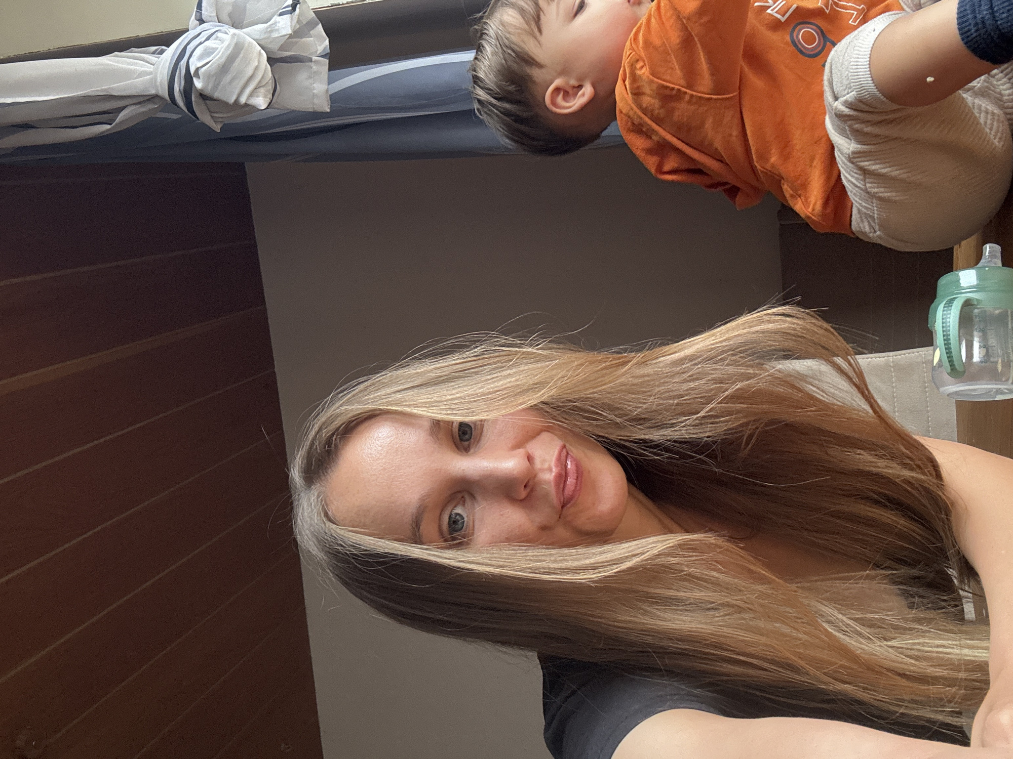 In a domestic setting, likely a room in Überherrn, Germany, a woman with long, blonde hair and blue eyes is looking into the camera, her mouth slightly pursed. She is wearing a black t-shirt. To her right, a young child wearing an orange hoodie and light-colored pants is visible from the side, facing away from the camera and appearing to be looking down. A light green baby bottle with a grey lid is partially visible in the foreground, to the left of the woman. The background features a wooden slatted ceiling and a window with a blue and white striped curtain. The lighting suggests it is daytime.