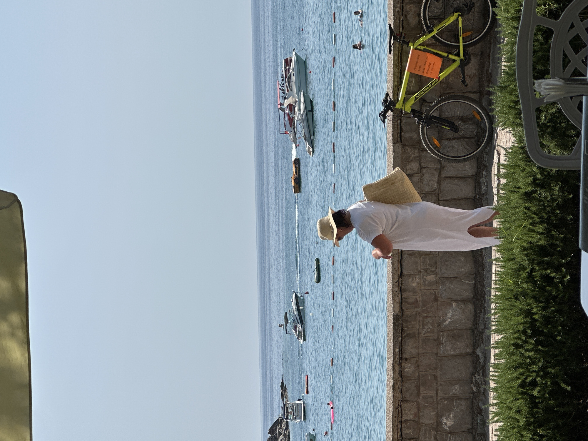 This image depicts a sunny day at the waterfront in Rafailovići, Montenegro. In the foreground, a woman wearing a white dress and a straw hat stands with her back to the camera, looking towards the sea and possibly checking her watch. She is carrying a straw bag over her shoulder. To her right, a bright yellow mountain bike is parked against a stone wall, with an orange sign attached to its handlebars. The sign appears to be an advertisement for a "Mountainbike for Sale" with a price and contact information in Italian.
The background features the calm, blue Adriatic Sea, dotted with various boats, including speedboats and smaller leisure craft. Several people are visible in the water, some swimming and others relaxing on inflatable rafts. A roped-off area demarcates a swimming zone. The coastline of Rafailovići, with its characteristic stone buildings and vegetation, is visible in the distance. The overall scene suggests a relaxed tourist atmosphere on a clear summer day.