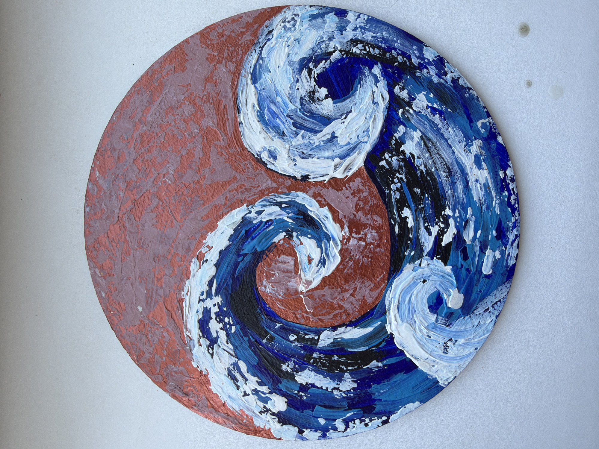 The image shows a circular painting of stylized waves against a reddish-brown textured background. The waves are depicted using thick, impasto strokes of blue and white paint, with hints of black in the darker areas. The overall style is abstract and dynamic, evoking a sense of movement and energy. The painting is placed on a white surface, possibly a windowsill or a table, with a plain white background. There are no people or specific landmarks visible that would indicate a location in Ufa, Russia.