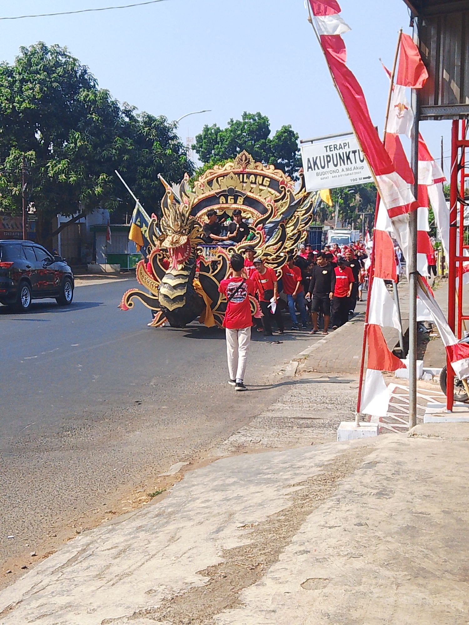 In Kalinyamatan, Indonesia, a parade features a large, ornate dragon float decorated with intricate golden carvings. The float is being pushed by a group of people dressed in red t-shirts and white pants. Some individuals in the group wear black shirts and shorts, also participating in the procession. A black SUV is visible on the left side of the road. Red and white flags line the right side of the street, interspersed with a sign that reads "TERAPI AKUPUNKTUR" and further details about a person named "BUDIYONO, A.Md. Akup." and an indication of "100 M" with an arrow. The event appears to be taking place outdoors on a sunny day, with trees and buildings in the background. The road is paved, and a sidewalk is present on the right side of the image.
