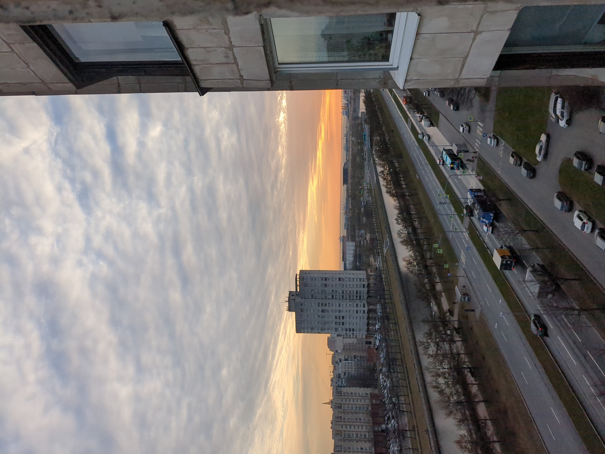 The image depicts an elevated view, likely from a high-rise building, overlooking a cityscape in Saint Petersburg, Russia. The sky is filled with clouds illuminated by the setting sun, casting a warm golden glow. A major thoroughfare, likely a multi-lane highway, is visible with cars and trucks moving along it. Rows of trees line the roadway, creating a park-like median. Buildings of varying heights, including a large apartment complex, stretch into the distance. Parked cars are visible on the sides of the road. A window frame from the building the image was taken from is visible in the top of the shot.