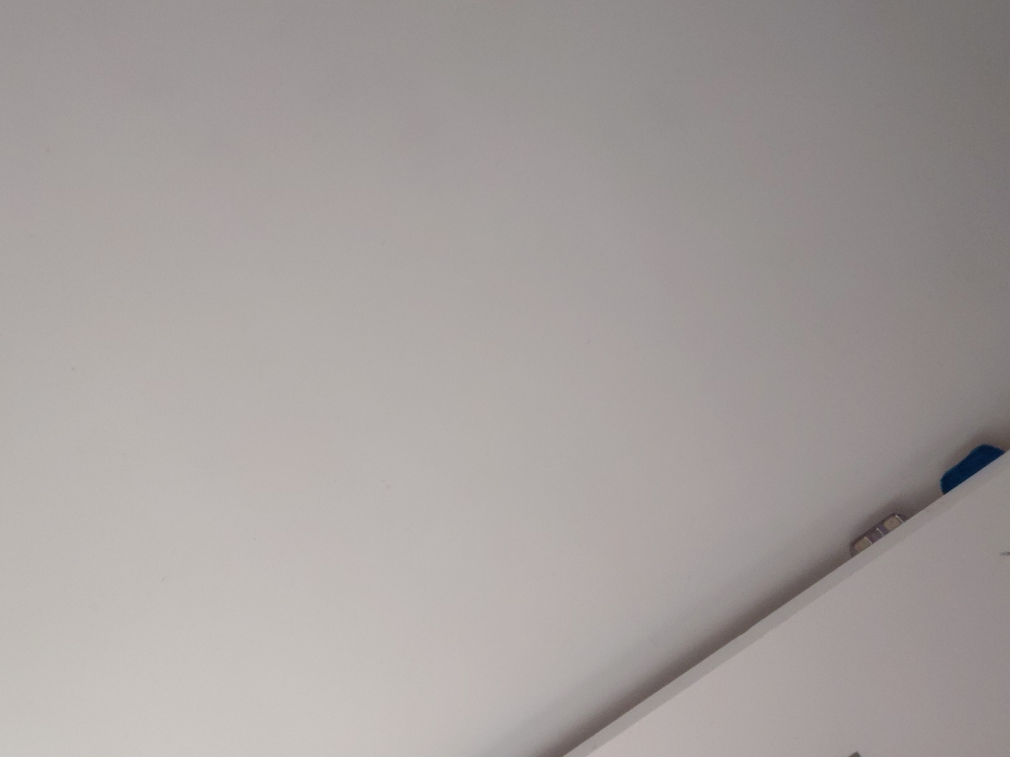 The image presents a close-up, static shot of an indoor architectural space, likely a ceiling or upper wall area in Santa Ana, Venezuela. The dominant subject is a vast, uniformly textured, light grey or off-white surface, which appears to be a painted wall or ceiling. A strong diagonal line cuts across the lower right portion of the frame, indicating an intersection with another light-colored surface, possibly a cabinet, shelf, or another wall. Along this diagonal edge, in the upper right quadrant of the image, several small, indistinct objects are visible. These include a small, dark blue object, possibly a piece of tape or a plastic component, located near the top right corner. Slightly to its left, there are two small, metallic or silver-colored objects, which could be clips, brackets, or small, recessed light fixtures, reflecting a subtle glint of light. No people are present, and no specific activity is taking place. The lighting is even and neutral, offering no cues about the time of day or outdoor weather. There is no visible text in the image. The scene is devoid of specific location identifiers beyond the provided context.