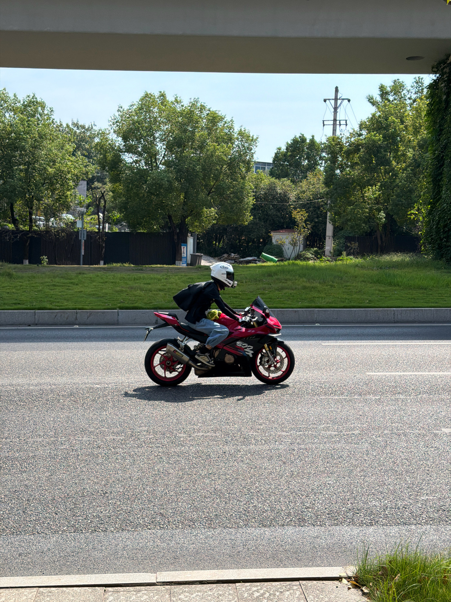 A person wearing a white helmet and a black jacket rides a red and black sport motorcycle on a paved road under a bright sky. The person is wearing blue jeans and white shoes, and a black backpack is visible on their back. The motorcycle has red wheels and a silver exhaust pipe. The road has white lane markings. In the background, there is a grassy area with trees and a power pole. The scene appears to be in Jiangjin, China, based on the visual cues typical of urban or suburban areas in the region.
